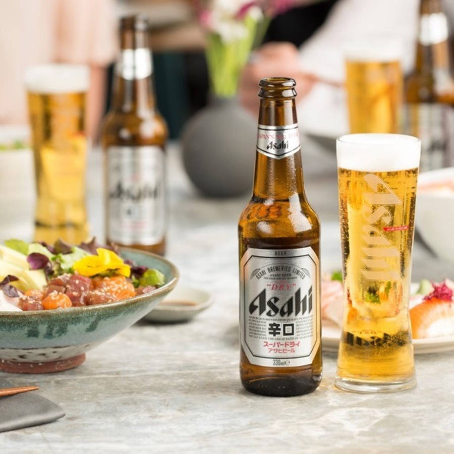 日本 桌布 壁紙 — 一瓶冰涼的Asahi Super Dry啤酒和玻璃杯放在大理石桌上，旁邊是一道色彩繽紛的日本料理。