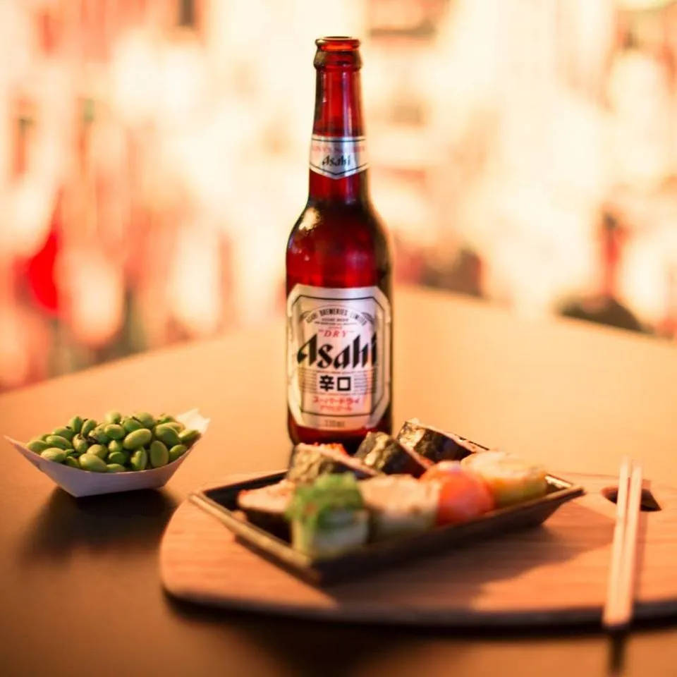 Beer 桌布 壁紙 — 一瓶冰涼的Asahi啤酒放在木托盤上，旁邊是一盤壽司和毛豆，在溫暖的環境光線中。