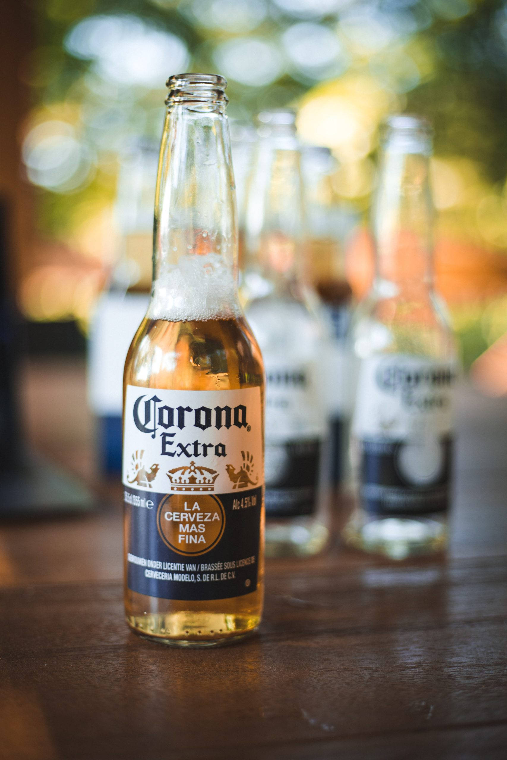 corona 桌布 壁紙 — 一瓶冰涼的Corona Extra啤酒瓶，帶有冷凝水珠，放在木桌上，背景有模糊的瓶子。