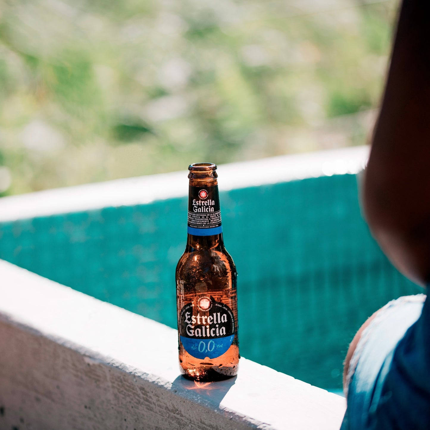 alcohol 桌布 壁紙 — 一瓶冰涼的Estrella Galicia 0.0啤酒放在陽光普照的泳池邊，旁邊有一位穿藍色衣服的人，背景有模糊的綠色樹葉。