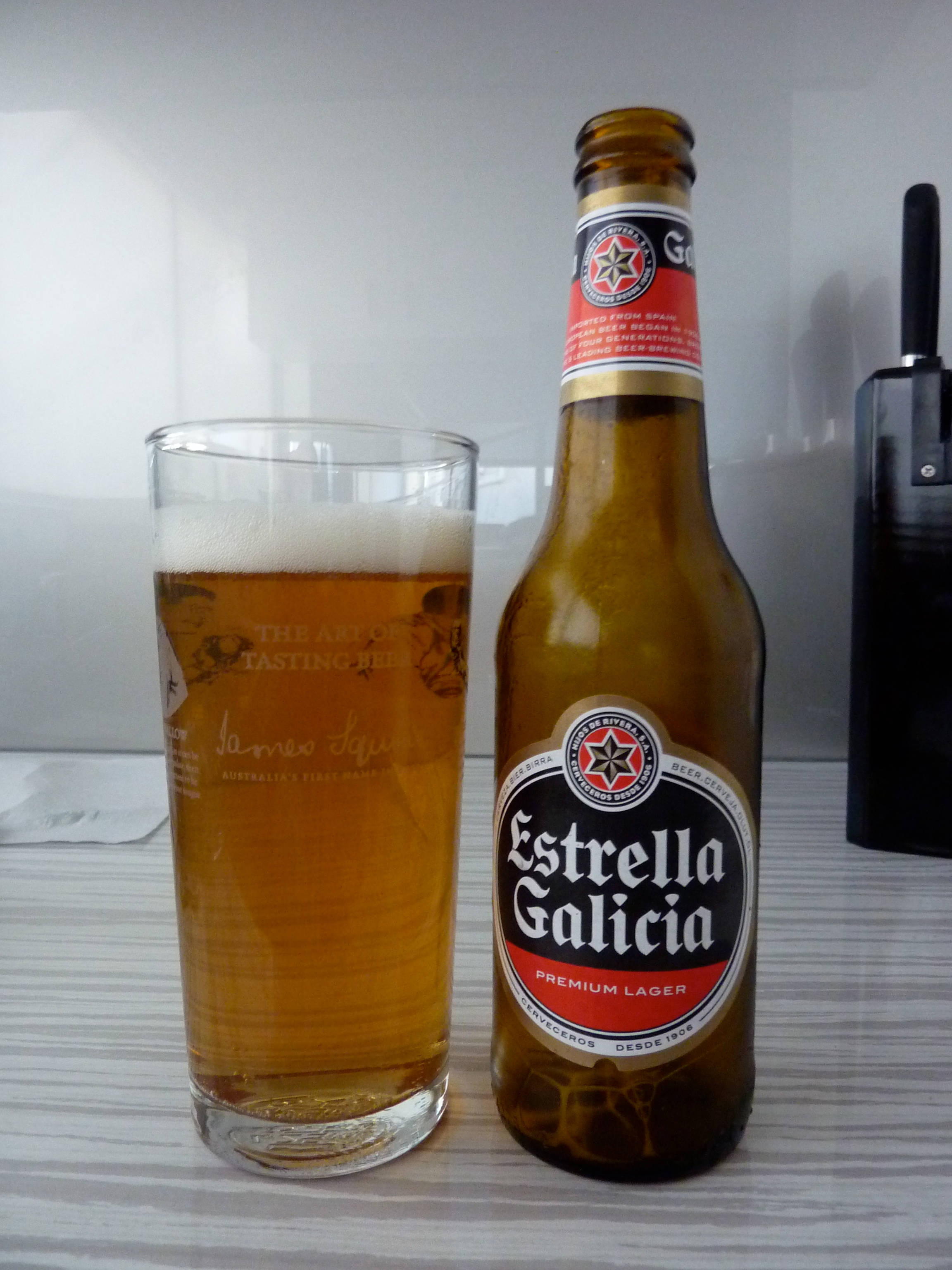 alcohol 桌布 壁紙 — 一瓶冰涼的Estrella Galicia啤酒放在淺色木桌上，旁邊有一杯滿滿的啤酒，頂部有泡沫。
