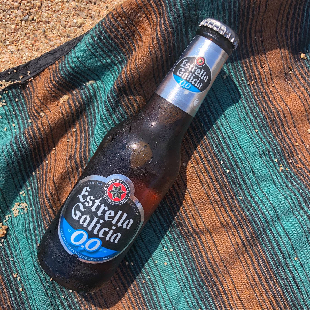 alcohol 桌布 壁紙 — 一瓶冰涼的Estrella Galicia 0.0啤酒瓶放在一條有沙粒散落的條紋毛巾上，在明亮的陽光下。