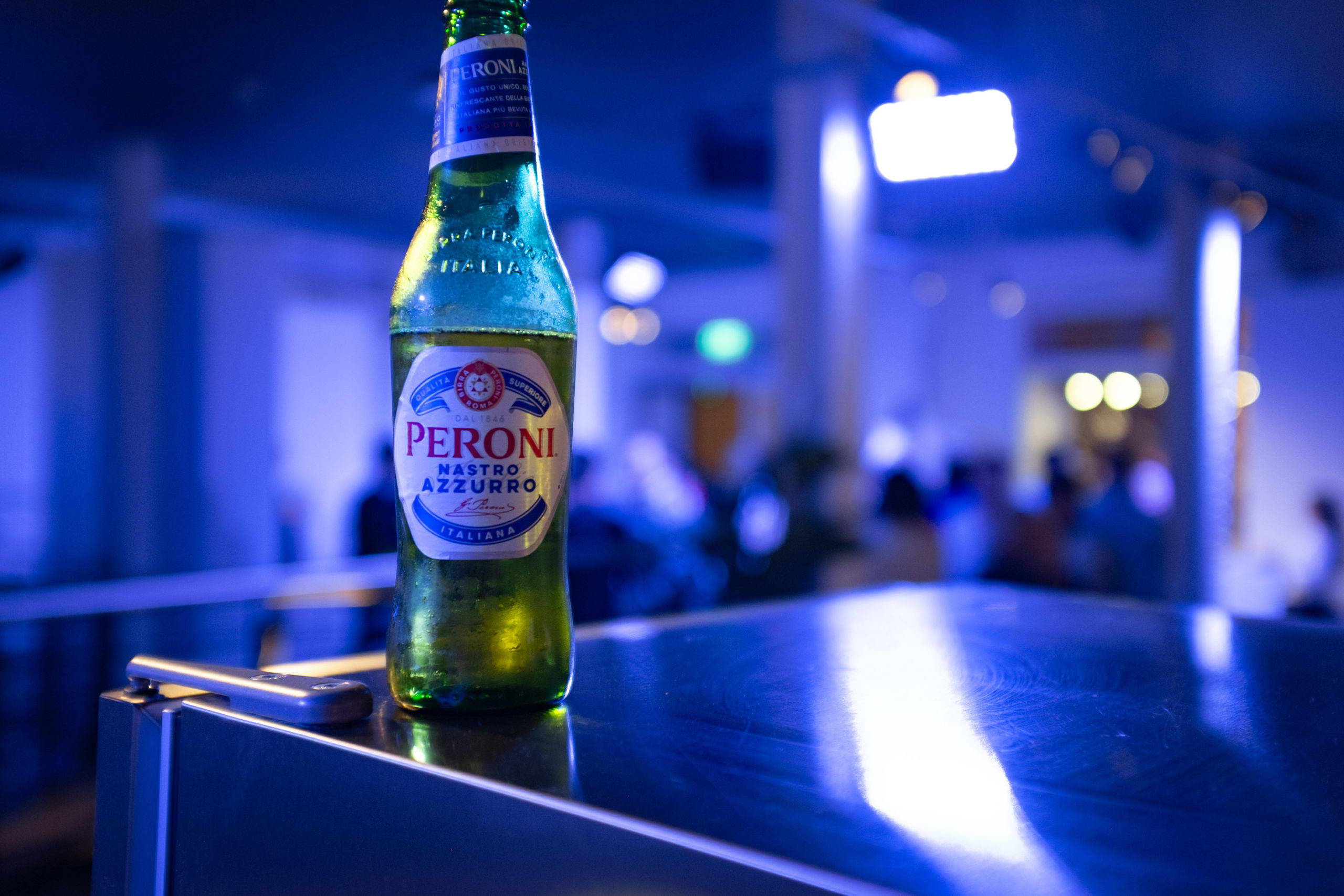 bottle 桌布 壁紙 — 一瓶冰涼的Peroni Nastro Azzurro啤酒在冷藍燈光下於反射的吧台上閃耀，背後是模糊的夜生活。