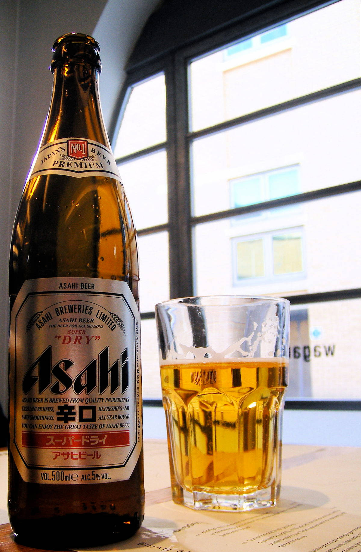 alcohol 桌布 壁紙 — 一瓶冰涼的朝日超乾啤酒和一個滿杯放在窗邊的桌子上，窗外可見城市建築。