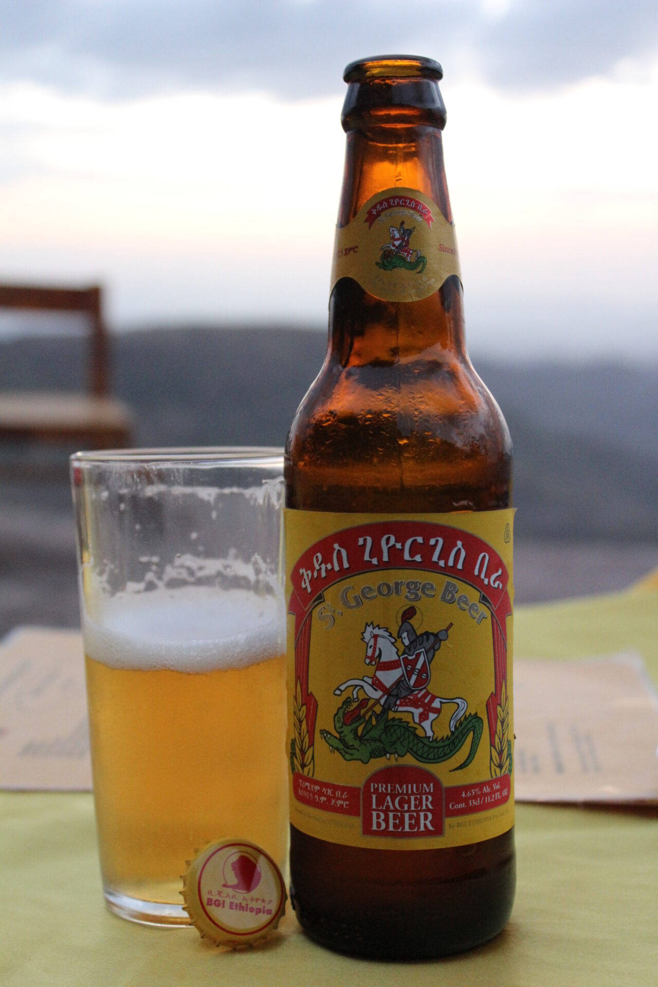 Beer 桌布 壁紙 — 一瓶冰涼的聖喬治啤酒和一個裝滿的杯子放在桌子上，俯瞰黃昏時霧氣瀰漫的海岸景色。
