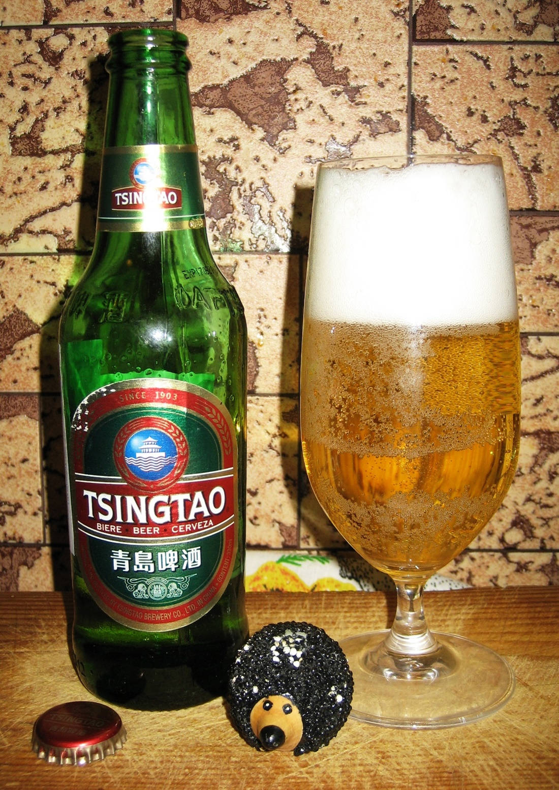booze 桌布 壁紙 — 一瓶冰涼的青島啤酒和一個有泡沫的玻璃杯放在木桌上，旁邊有一個刺蝟公仔，背景是磁磚牆。
