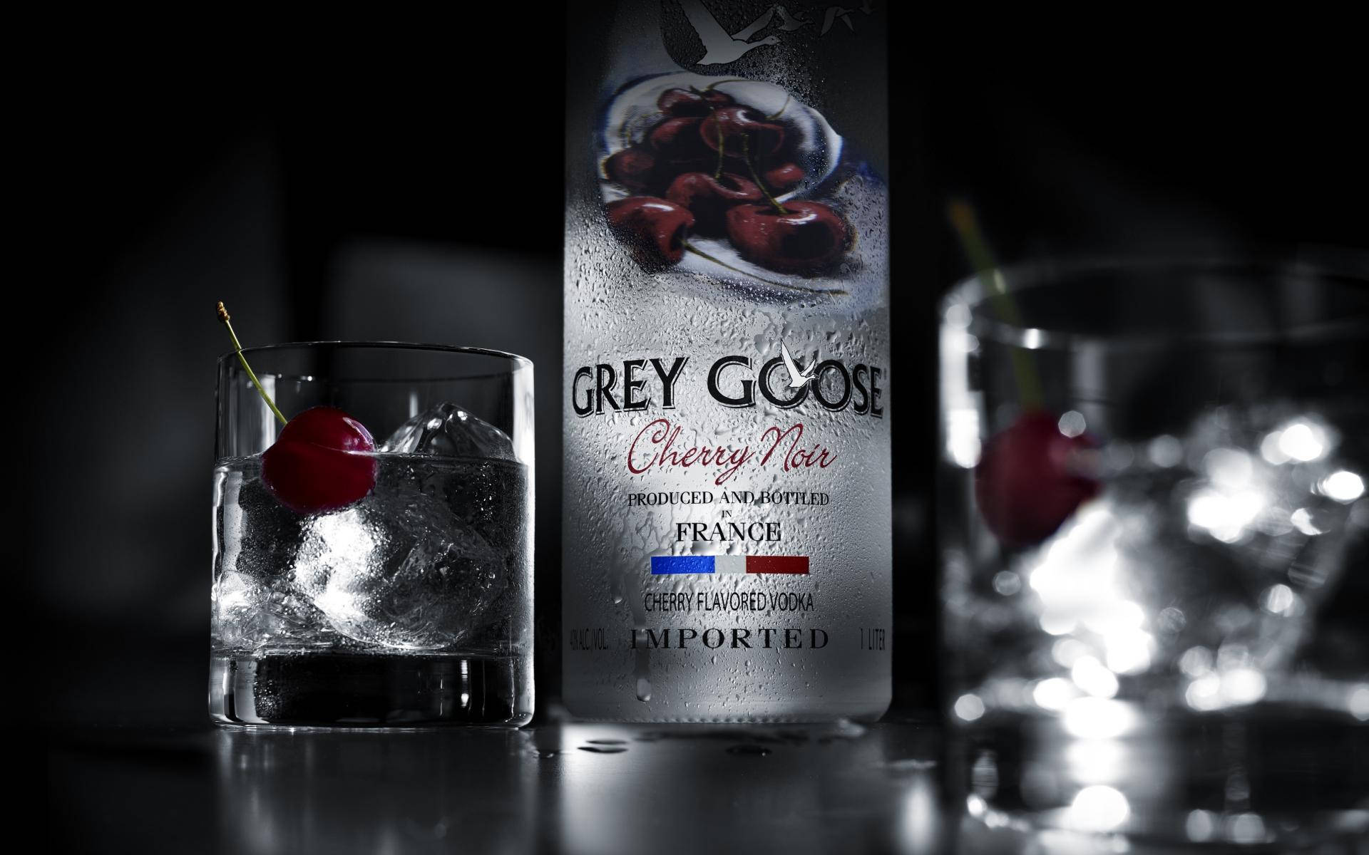 飲料 桌布 壁紙 — 一瓶冰鎮的 Grey Goose Cherry Noir 伏特加，搭配冰塊和櫻桃裝飾，置於深色反光表面上的玻璃杯中。
