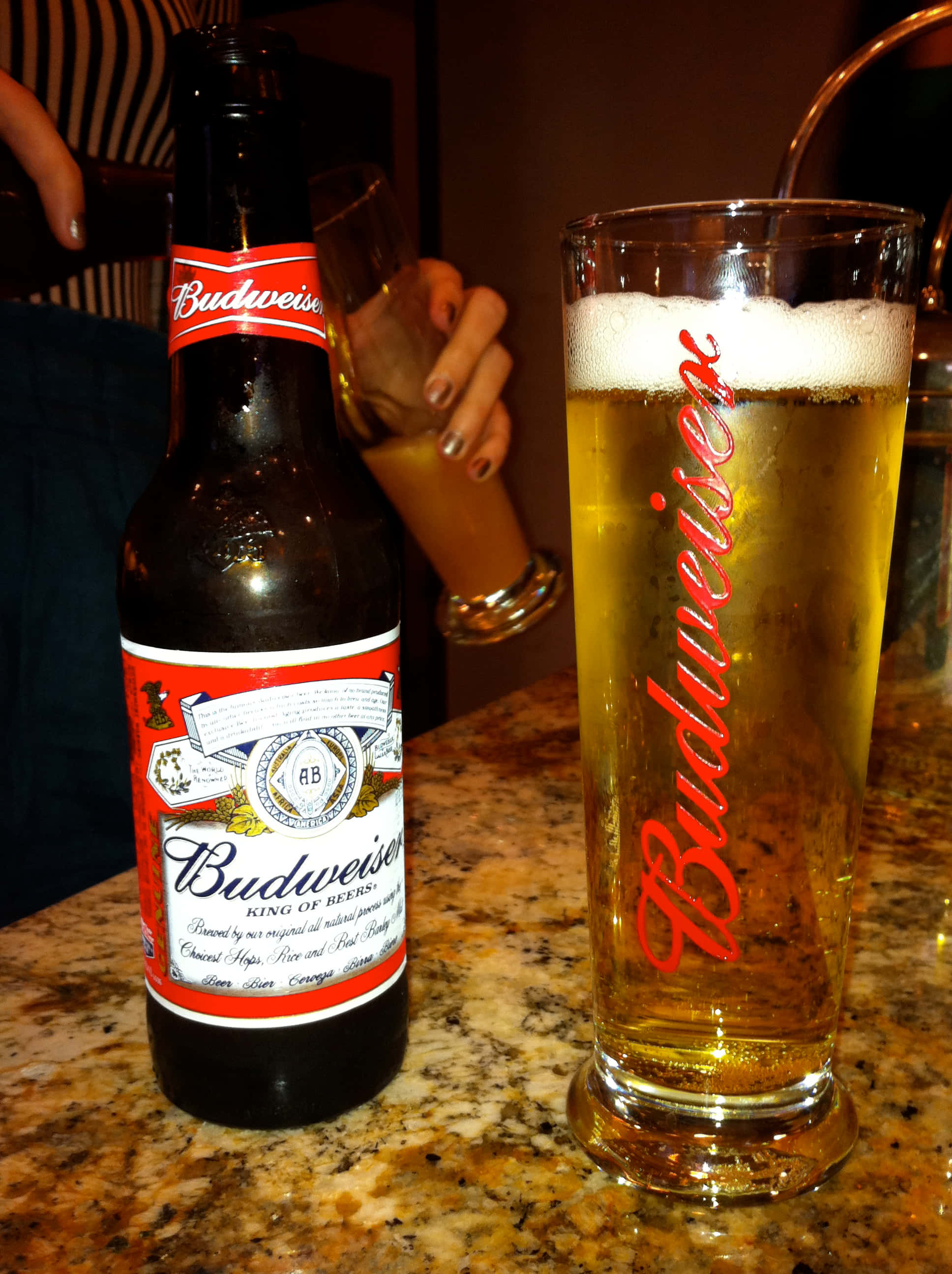 Beer 桌布 壁紙 — 一瓶冰鎮的Budweiser啤酒和一個玻璃杯放在有斑點的酒吧櫃檯上，背景中有一隻手拿著另一個玻璃杯。