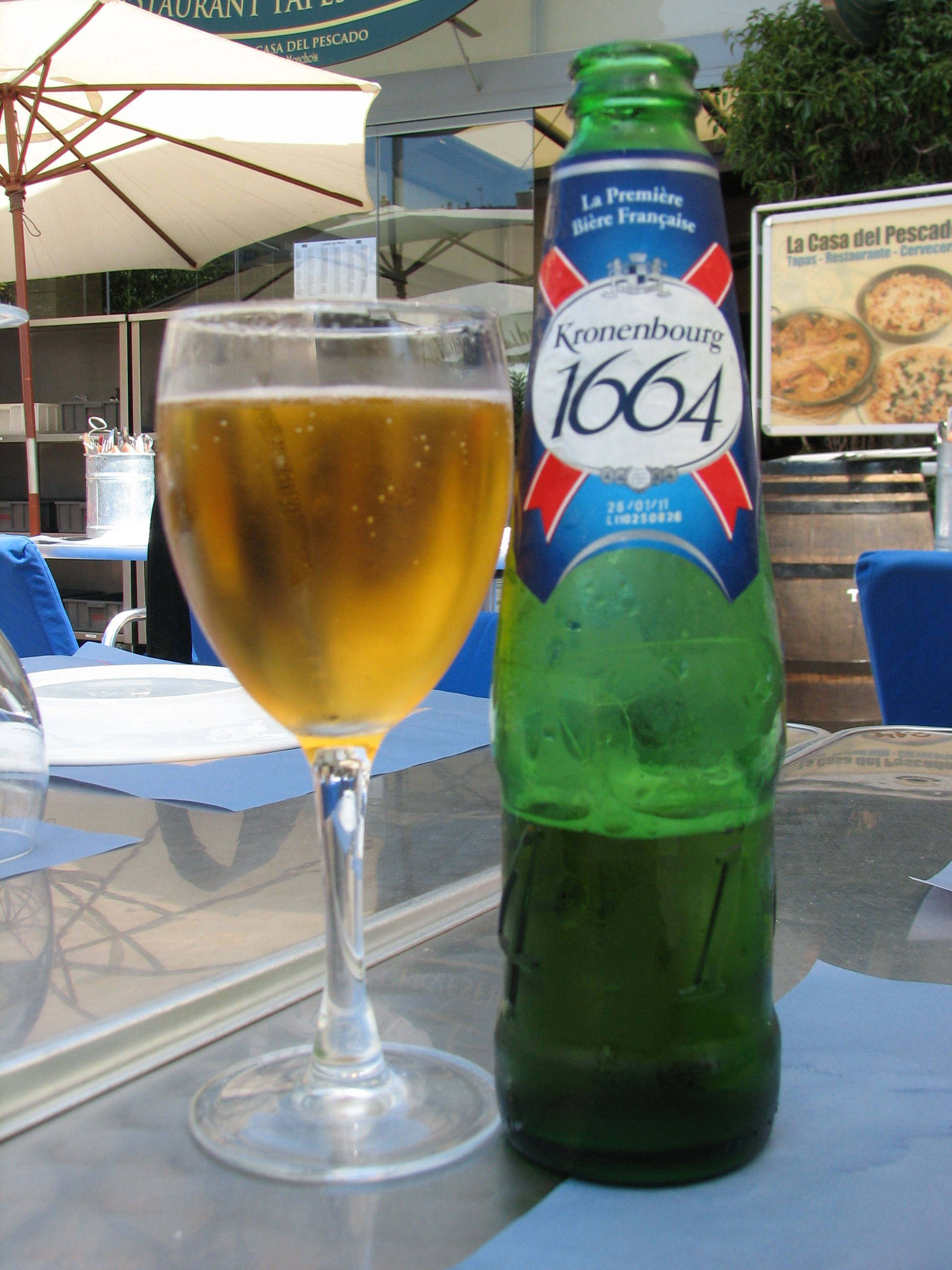 Outdoor 桌布 壁紙 — 一瓶冰鎮的Kronenbourg 1664啤酒瓶和玻璃杯放在白色遮陽傘下的戶外咖啡廳玻璃桌上。