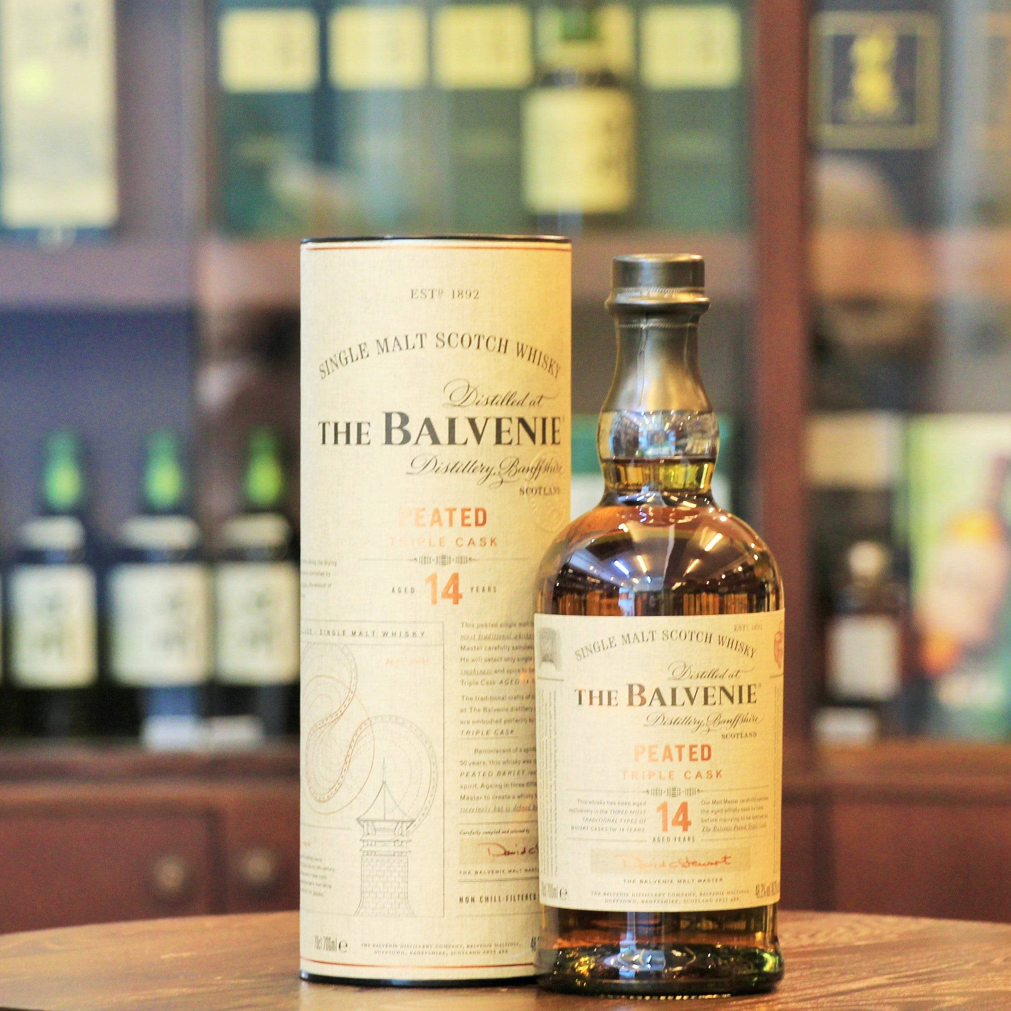 飲料 桌布 壁紙 — 一瓶及一盒The Balvenie 14年陳釀泥煤三重桶單一麥芽威士忌放在木製吧檯上，背景有模糊的酒瓶。