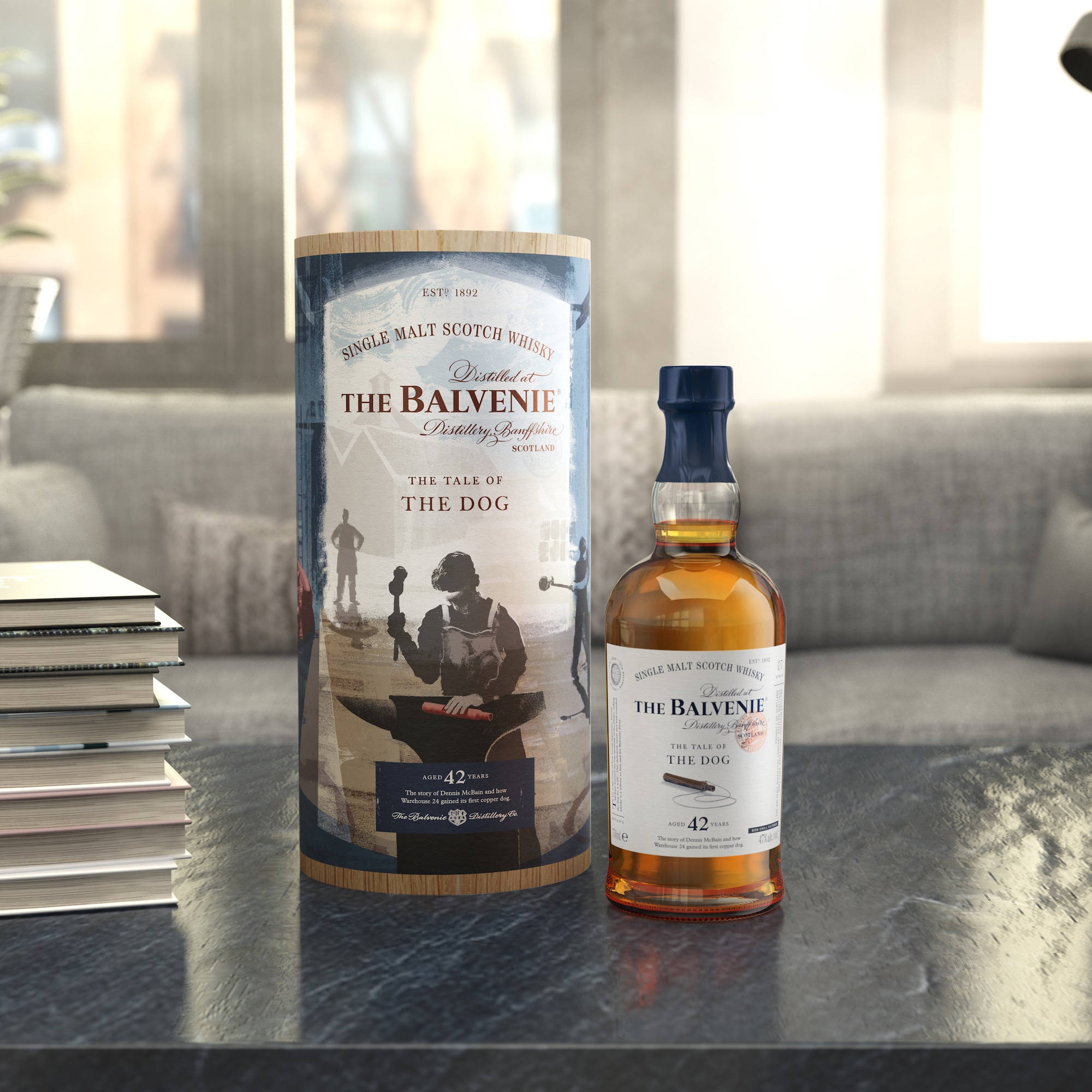 Drink 桌布 壁紙 — 一瓶和一個管裝的 The Balvenie 42 Year Old 威士忌放在一張深色桌子上，旁邊有一堆書，背景是灰色沙發和明亮的窗戶。