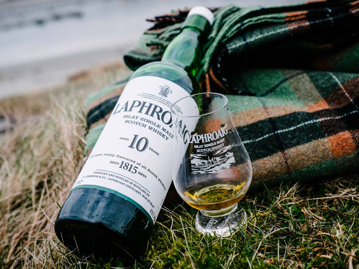 iPhone 桌布 壁紙 — 一瓶和一杯Laphroaig 10年艾雷島單一麥芽威士忌放在海灘附近的草地上，旁邊有一條格子毯。