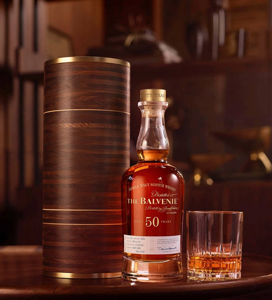 飲料 桌布 壁紙 — 一瓶奢華的50年Balvenie威士忌，連同木管和酒杯，放置在深色木質表面上，周圍是溫暖的環境燈光。