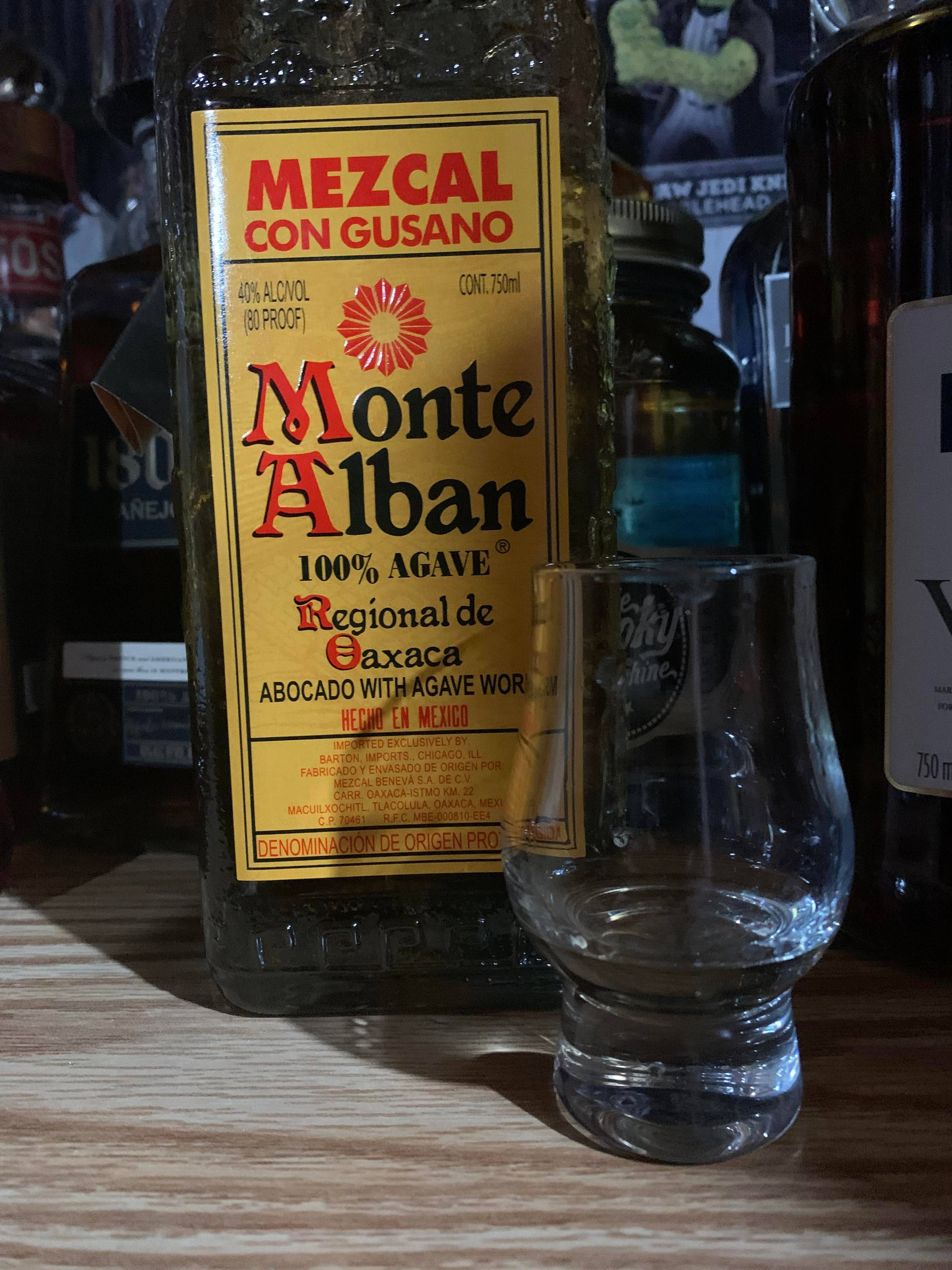 Drink 桌布 壁紙 — 一瓶帶有gusano的Monte Alban梅斯卡爾酒放在木製吧檯上，旁邊有一個空的品酒杯。