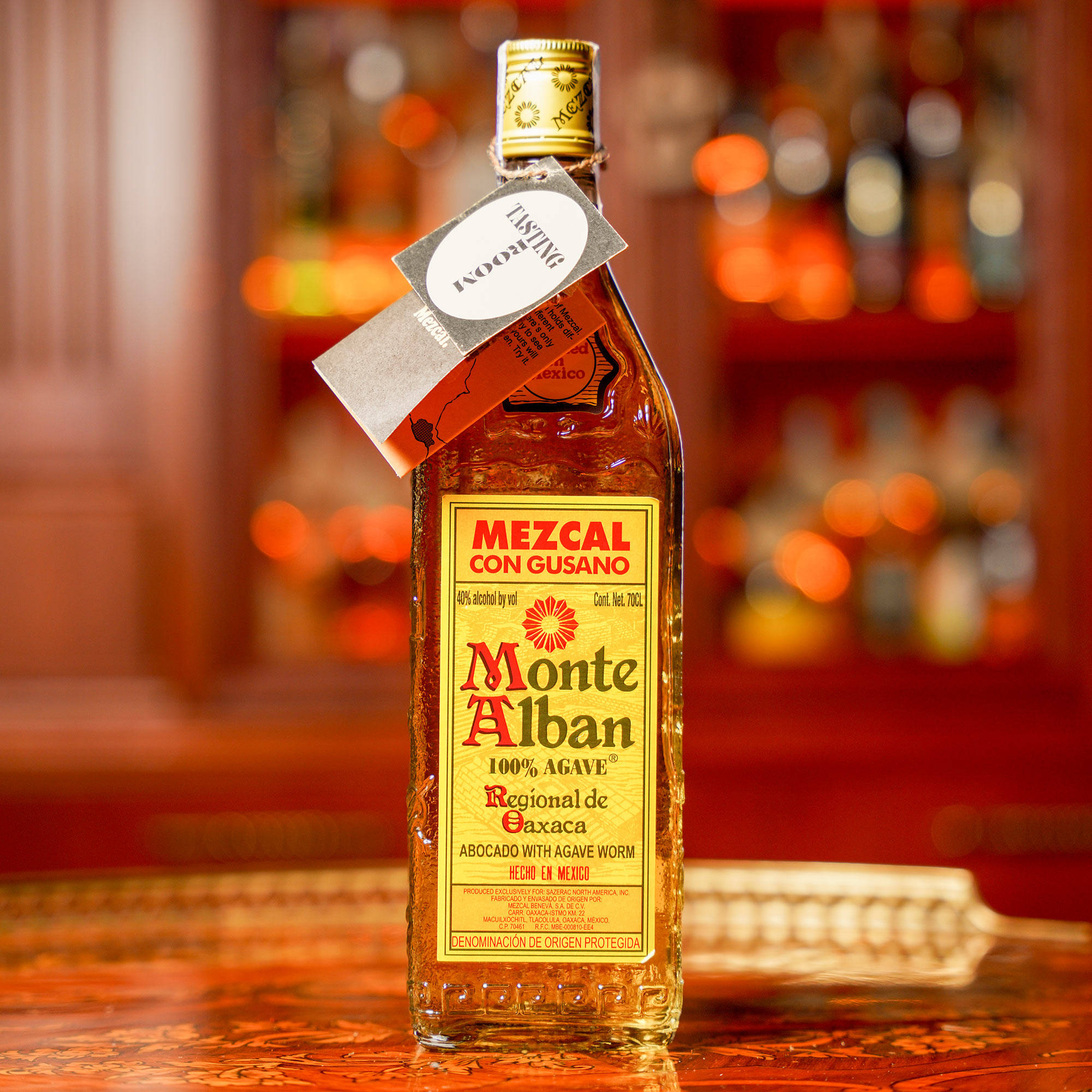 Drink 桌布 壁紙 — 一瓶帶有蟲子的Monte Alban梅斯卡爾酒放在有圖案的吧檯上，背景是溫暖的散景燈光。