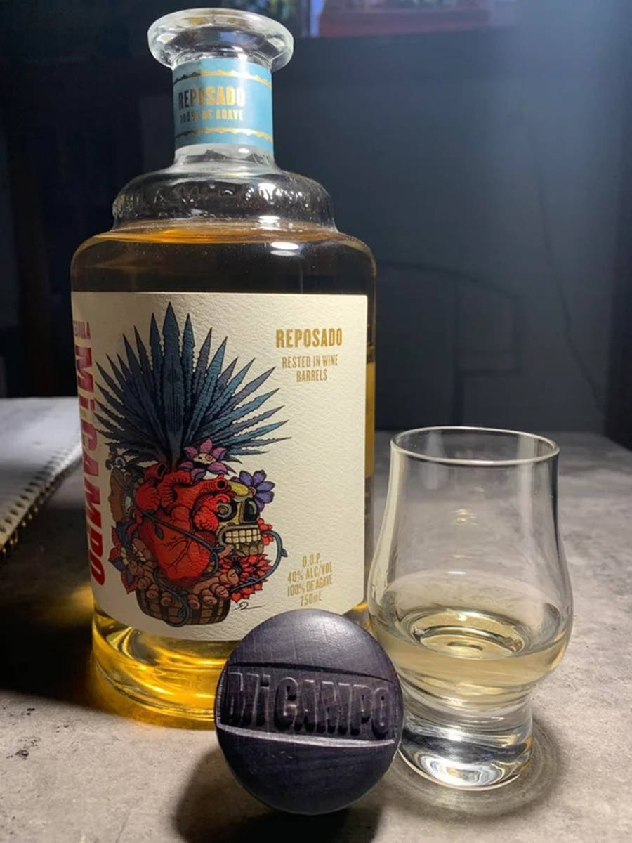 glass 桌布 壁紙 — 一瓶帶有骷髏標籤的Mi Campo reposado龍舌蘭酒，旁邊放著一個品酒杯和一塊品牌石頭，置於石檯面上。