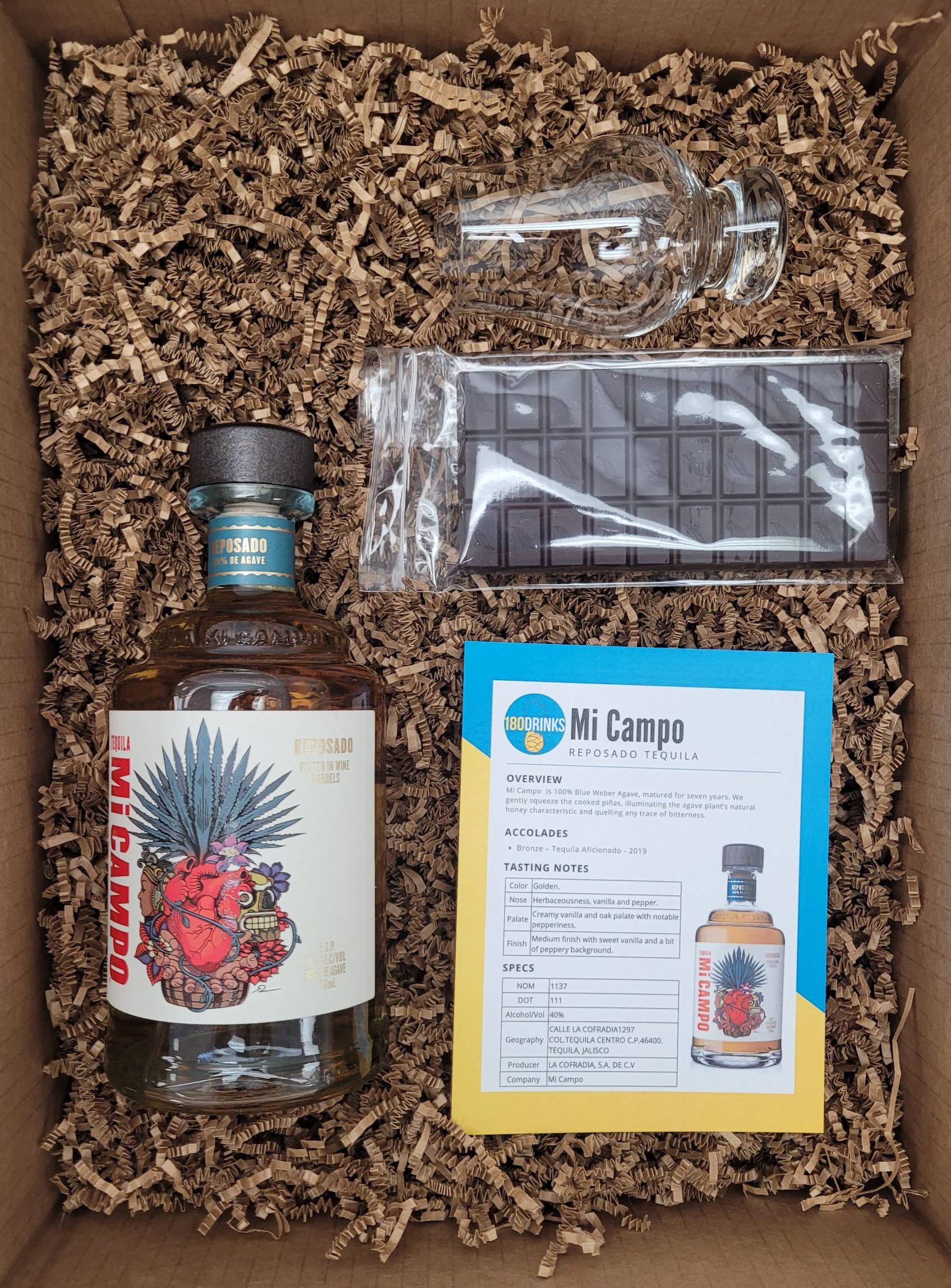 Box 桌布 壁紙 — 一瓶帶有鮮豔標籤的Mi Campo reposado龍舌蘭酒，放在一個裝滿碎棕色紙板的紙箱中，旁邊還有一條包裝好的巧克力條和一張資訊卡。