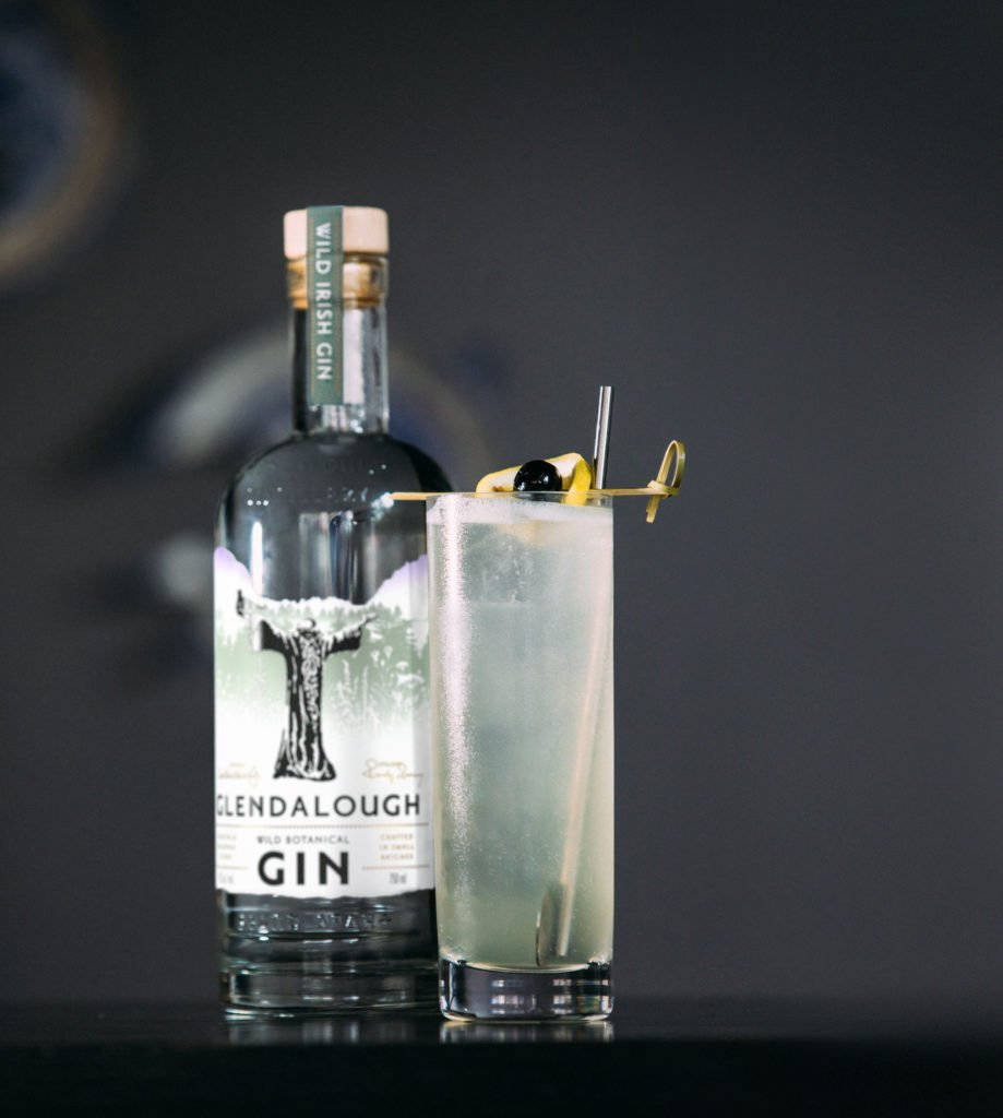 Gin 桌布 壁紙 — 一瓶時尚的Glendalough Wild Irish Gin旁邊放著一杯高身琴酒雞尾酒，飾以檸檬扭紋和橄欖。