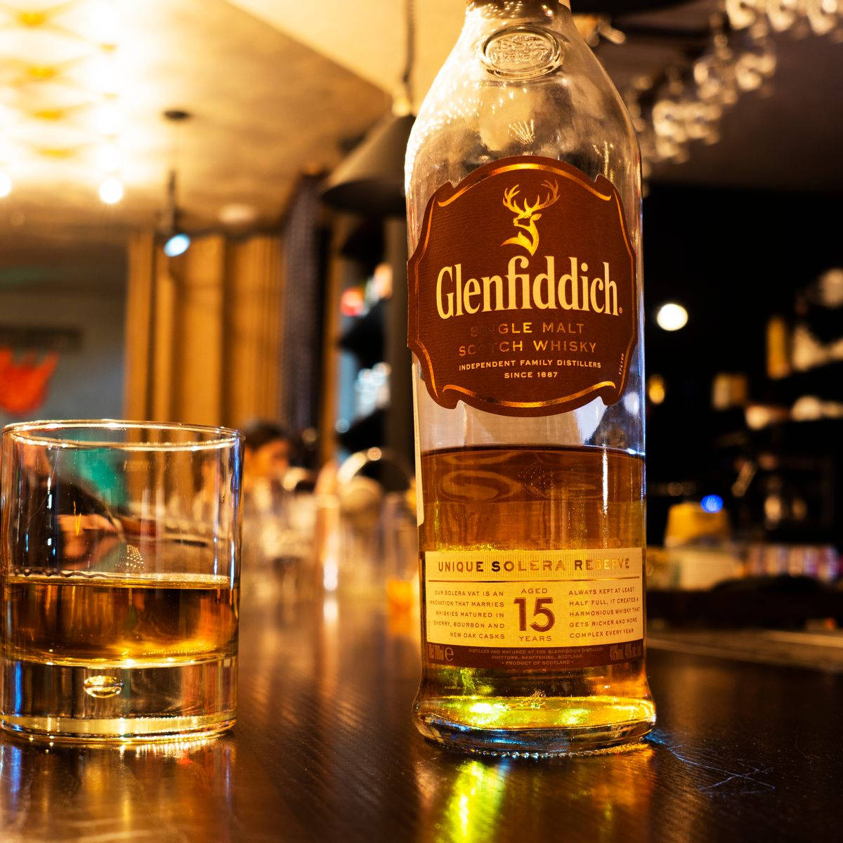 alcohol 桌布 壁紙 — 一瓶格蘭菲迪15年威士忌和一個玻璃杯放在拋光吧檯上，溫暖的環境燈光下。