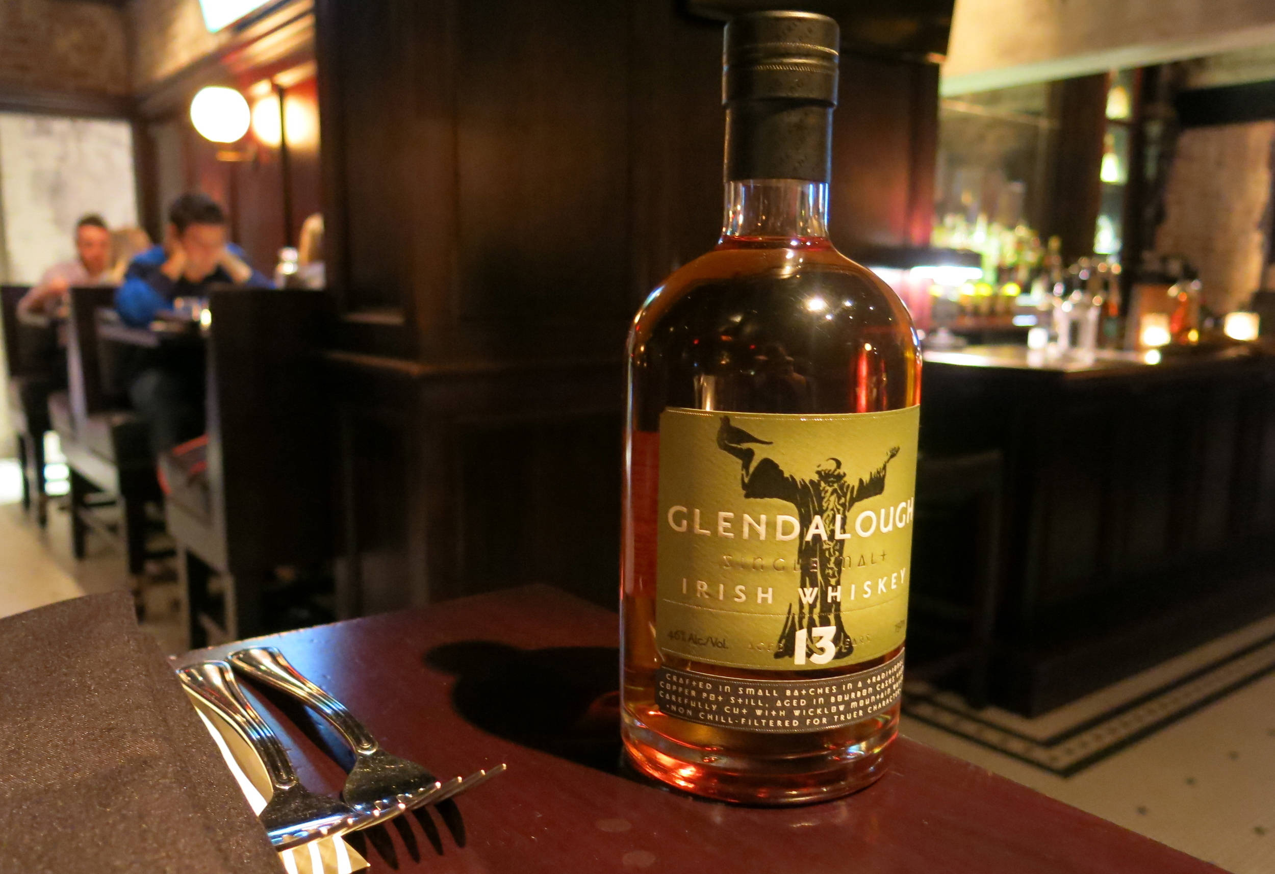 Whiskey 桌布 壁紙 — 一瓶格蘭達洛愛爾蘭威士忌在昏暗酒吧的木桌上溫暖地發光，背景有顧客。