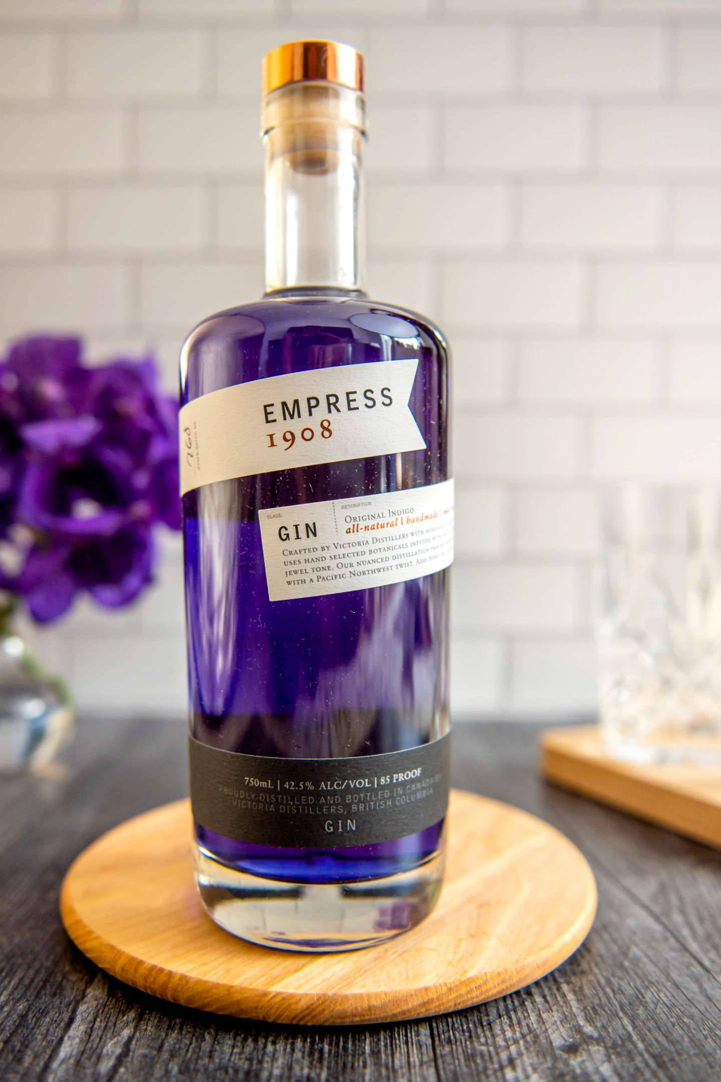 bottle 桌布 壁紙 — 一瓶標有 Empress 1908 的時尚紫色琴酒放在木杯墊上，背景是白色瓷磚牆，附近有紫色花朵。