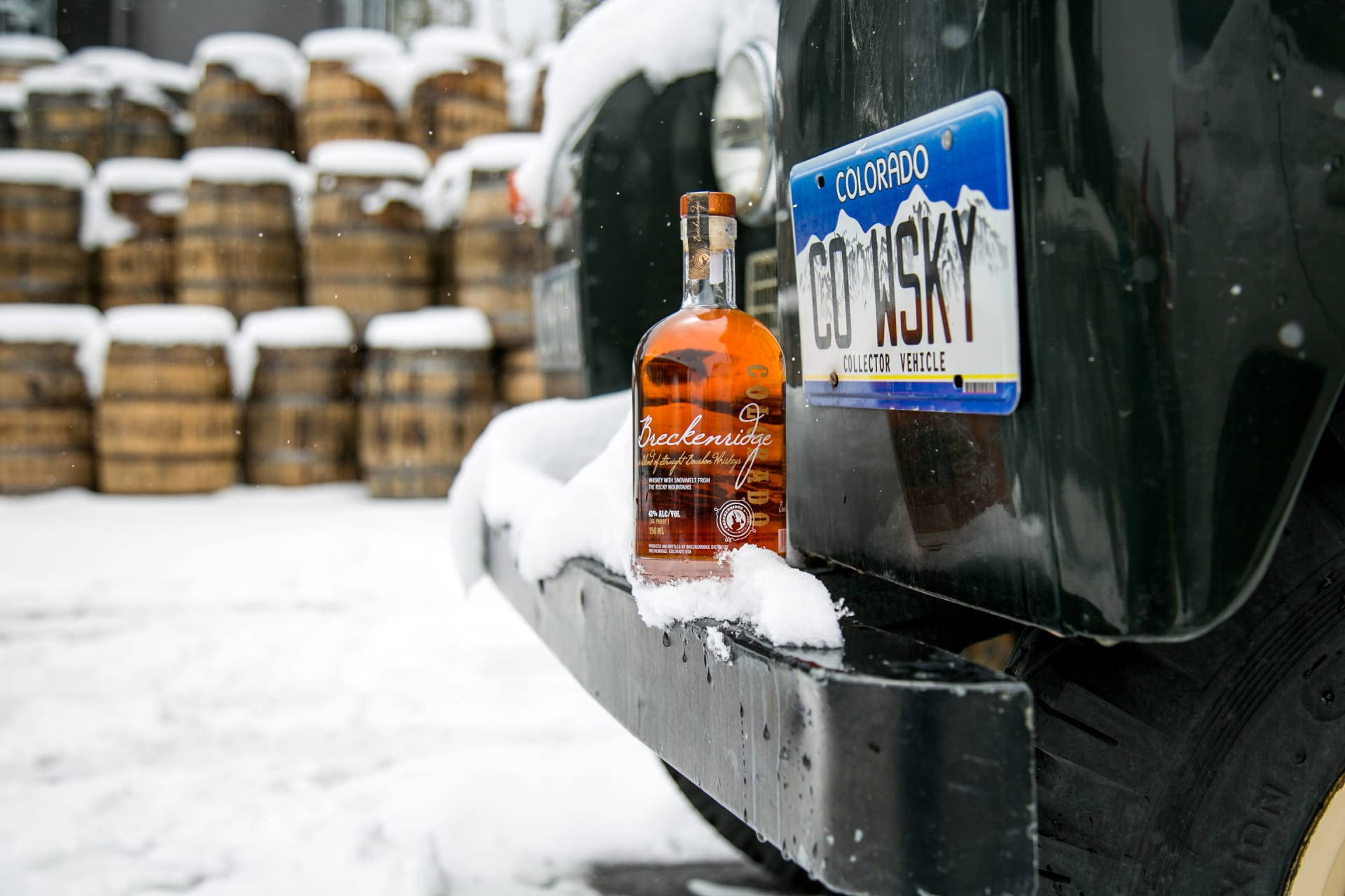 Drink 桌布 壁紙 — 一瓶波本威士忌放在黑色卡車的保險桿上的雪中，旁邊是印有「CO WSKY」的科羅拉多州車牌，背景是雪中的威士忌酒桶。