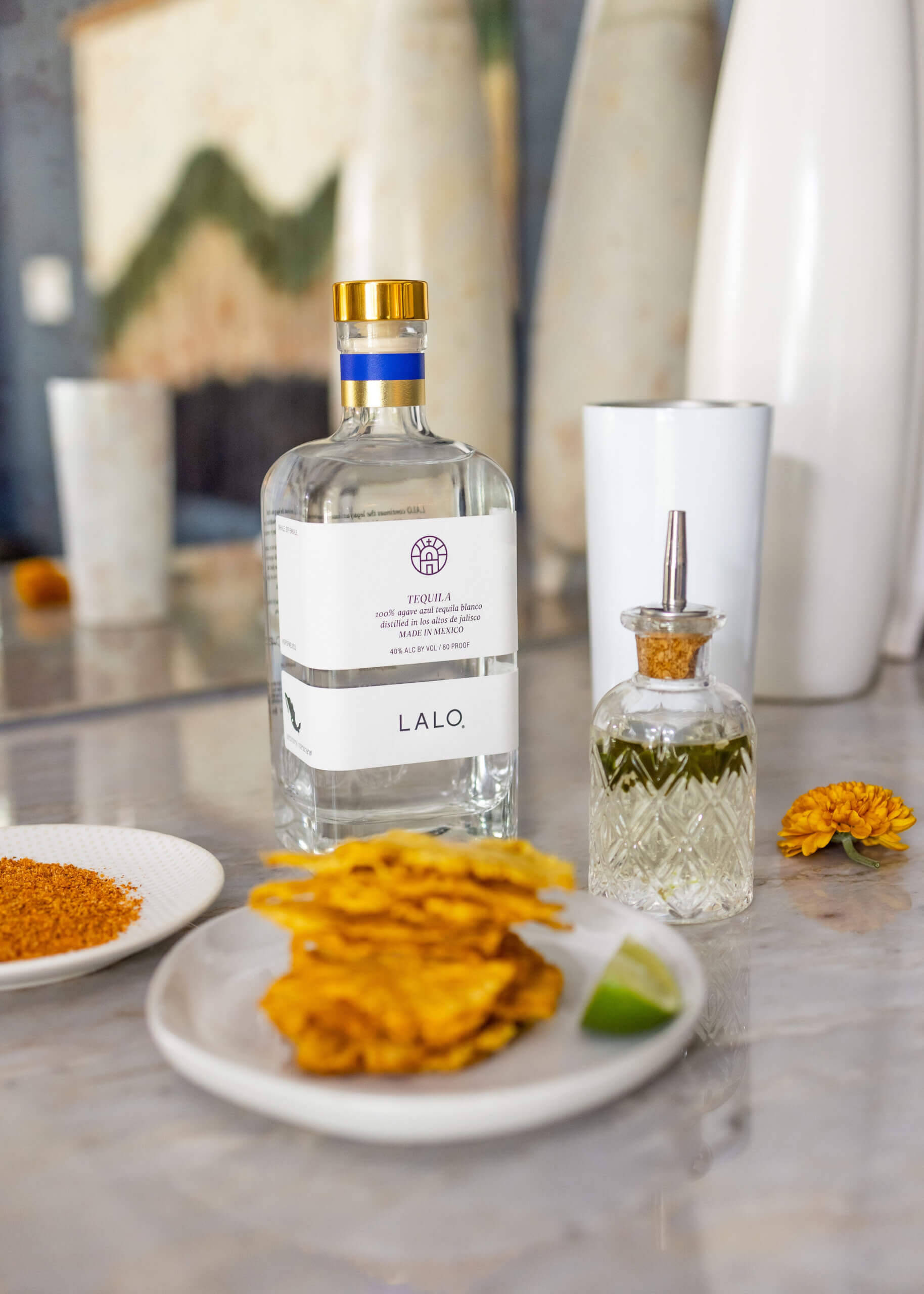 alcohol 桌布 壁紙 — 一瓶流線型的Lalo龍舌蘭酒放在大理石檯面上，旁邊有脆薯片、萊姆和花卉裝飾。