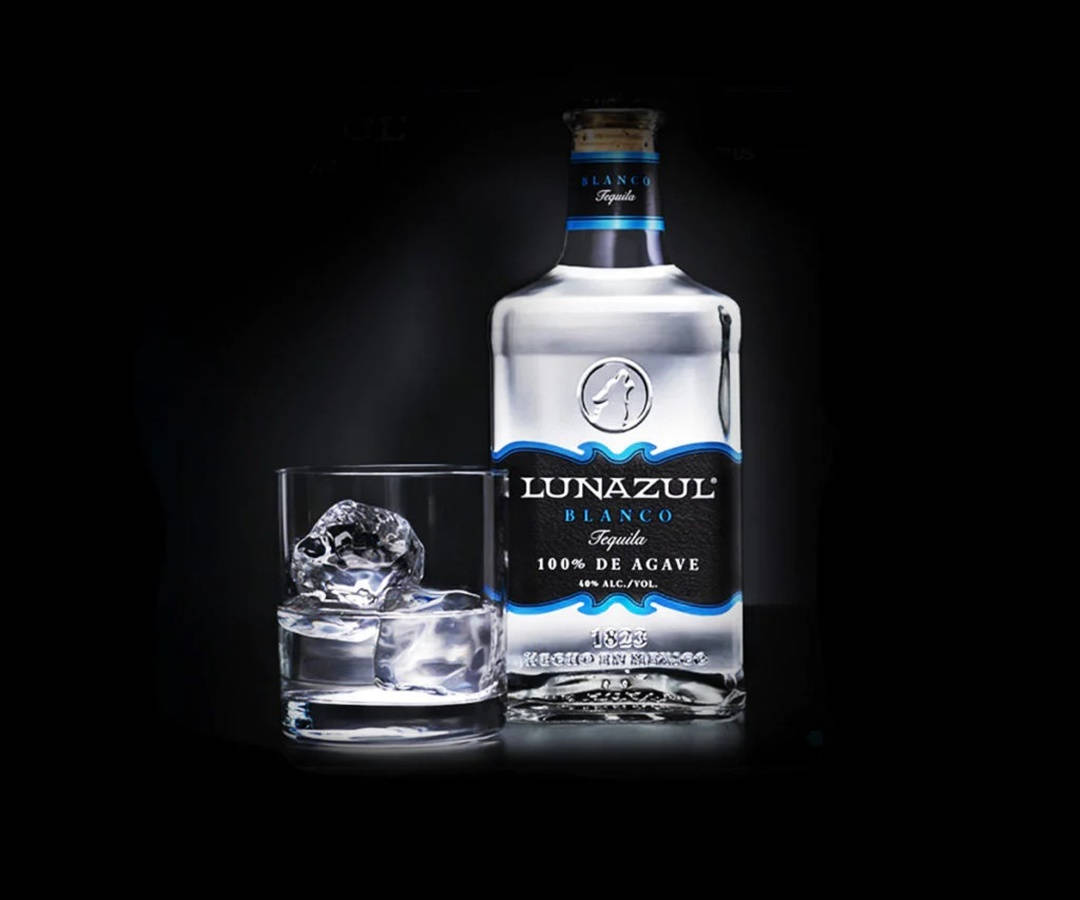 alcohol 桌布 壁紙 — 一瓶流線型的Lunazul Blanco龍舌蘭酒放在一個裝有大冰塊的玻璃杯旁邊，背景為深色。