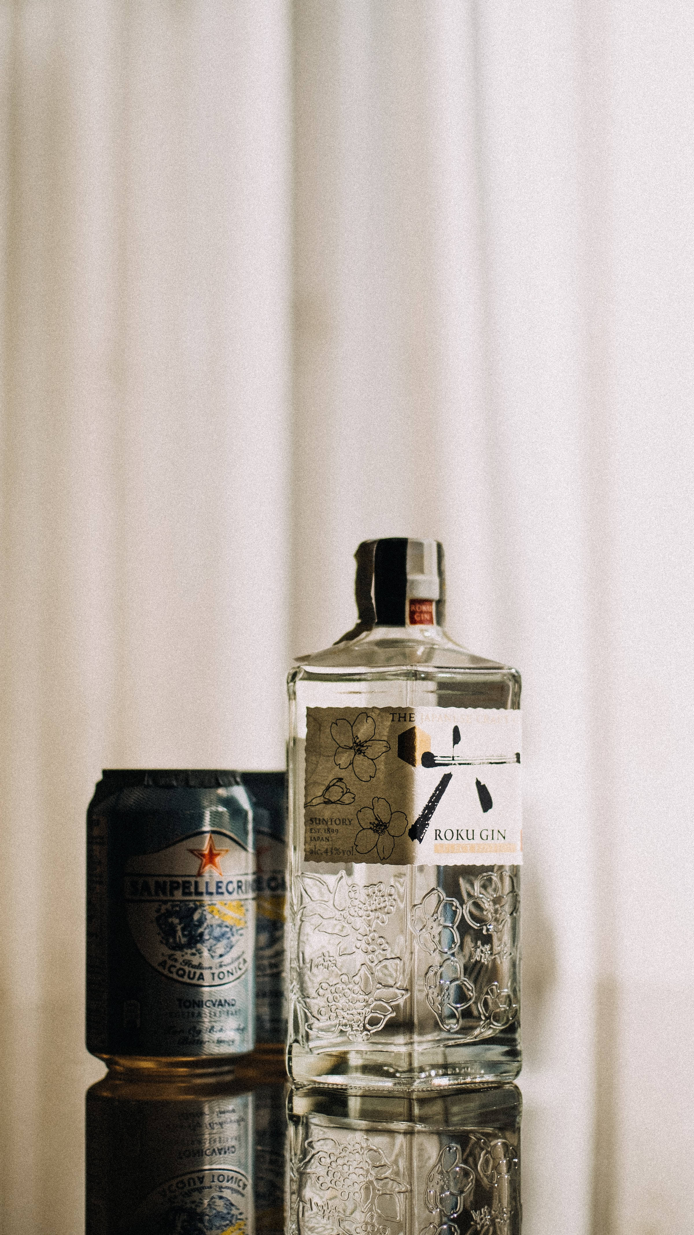 drinks 桌布 壁紙 — 一瓶流線型的Roku Gin和San Pellegrino tonic優雅地放置在反光表面上，背景是柔軟的白色窗簾。