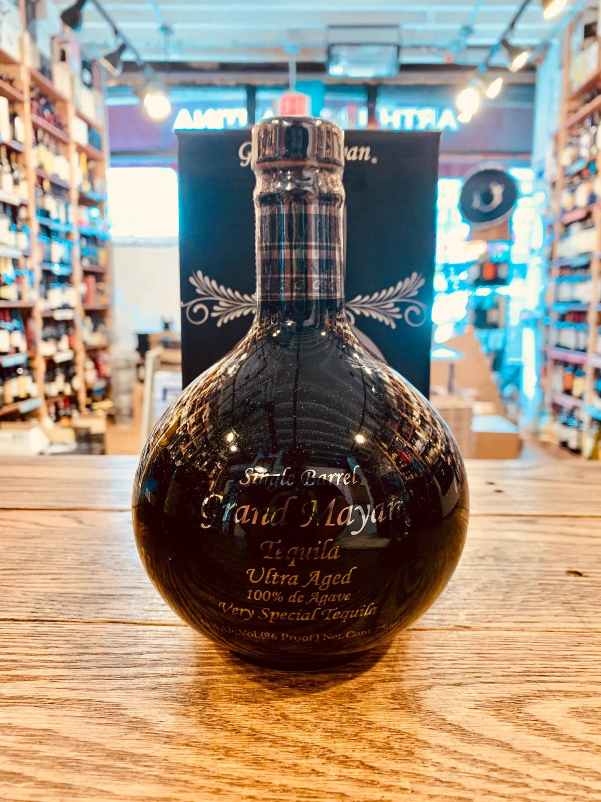 alcohol 桌布 壁紙 — 一瓶深色玻璃瓶裝的 Grand Mayan Ultra Aged Tequila 置中放在木製櫃檯上，背景是溫暖燈光的酒舖，架上陳列著各種酒瓶。