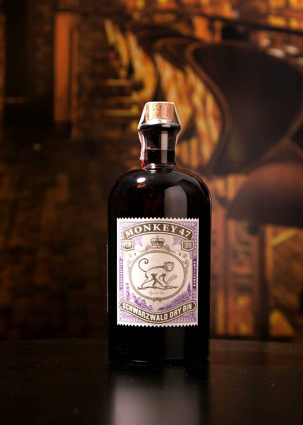 Drink 桌布 壁紙 — 一瓶深色玻璃瓶裝的Monkey 47琴酒，帶有紫色標籤，上面有猴子插圖，放置在反光表面上，背景是溫暖模糊的木質背景。