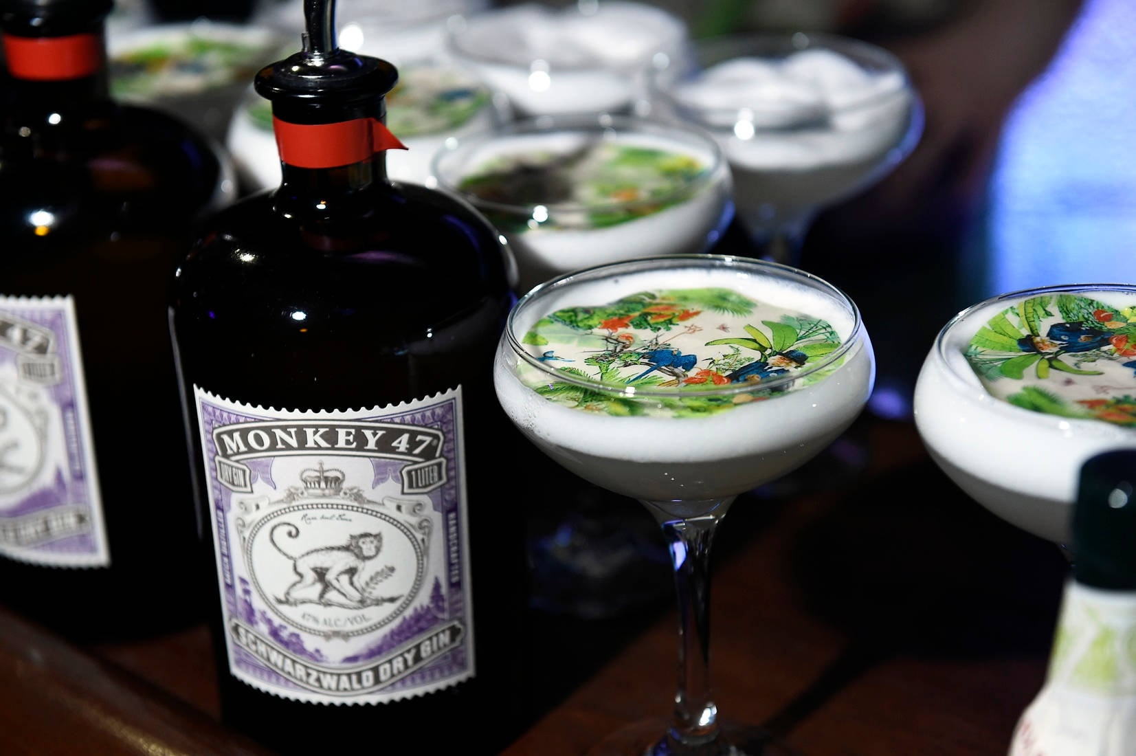 cocktail 桌布 壁紙 — 一瓶深色的Monkey 47琴酒放在優雅的庫佩酒杯旁，杯中盛滿了奶油雞尾酒，上面點綴著充滿活力的叢林主題可食用藝術品。