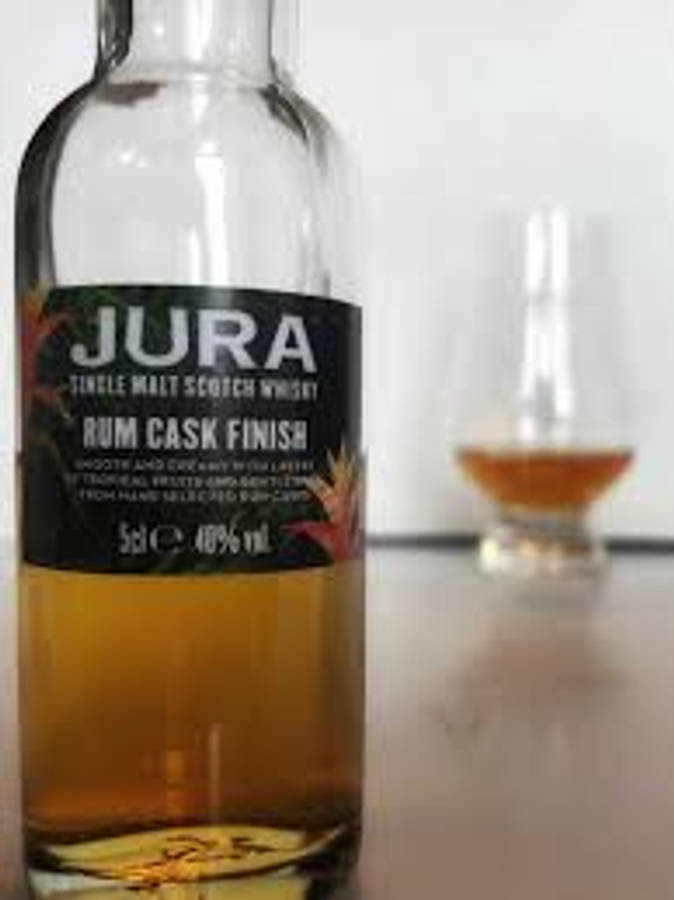 安卓 桌布 壁紙 — 一瓶清晰的Jura Rum Cask Finish威士忌在焦點中，背景中有一杯琥珀色液體模糊不清。