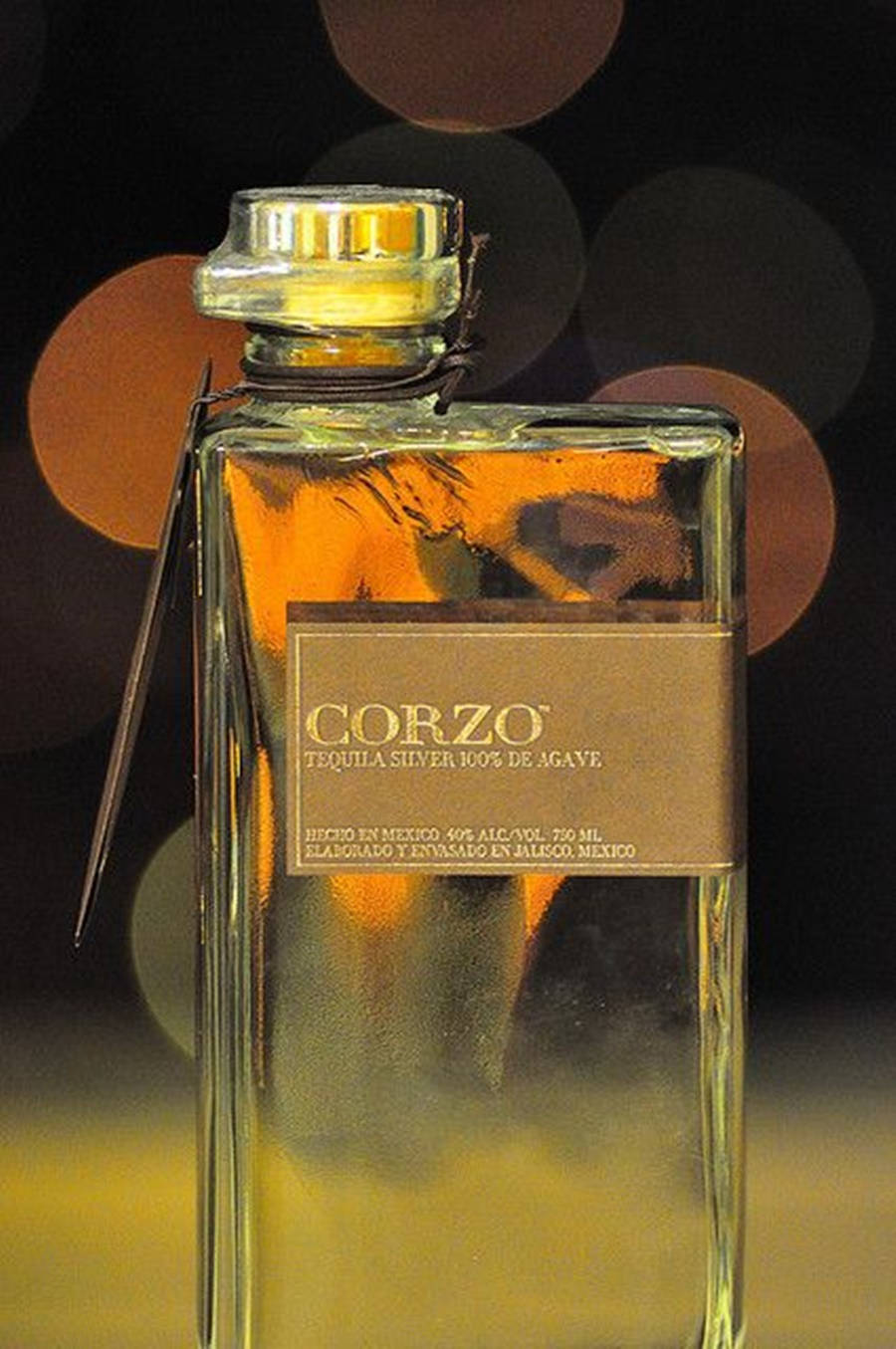 bottle 桌布 壁紙 — 一瓶清澈的 Corzo Silver Tequila 玻璃瓶，在溫暖的散景燈光背景下閃耀著金色液體。