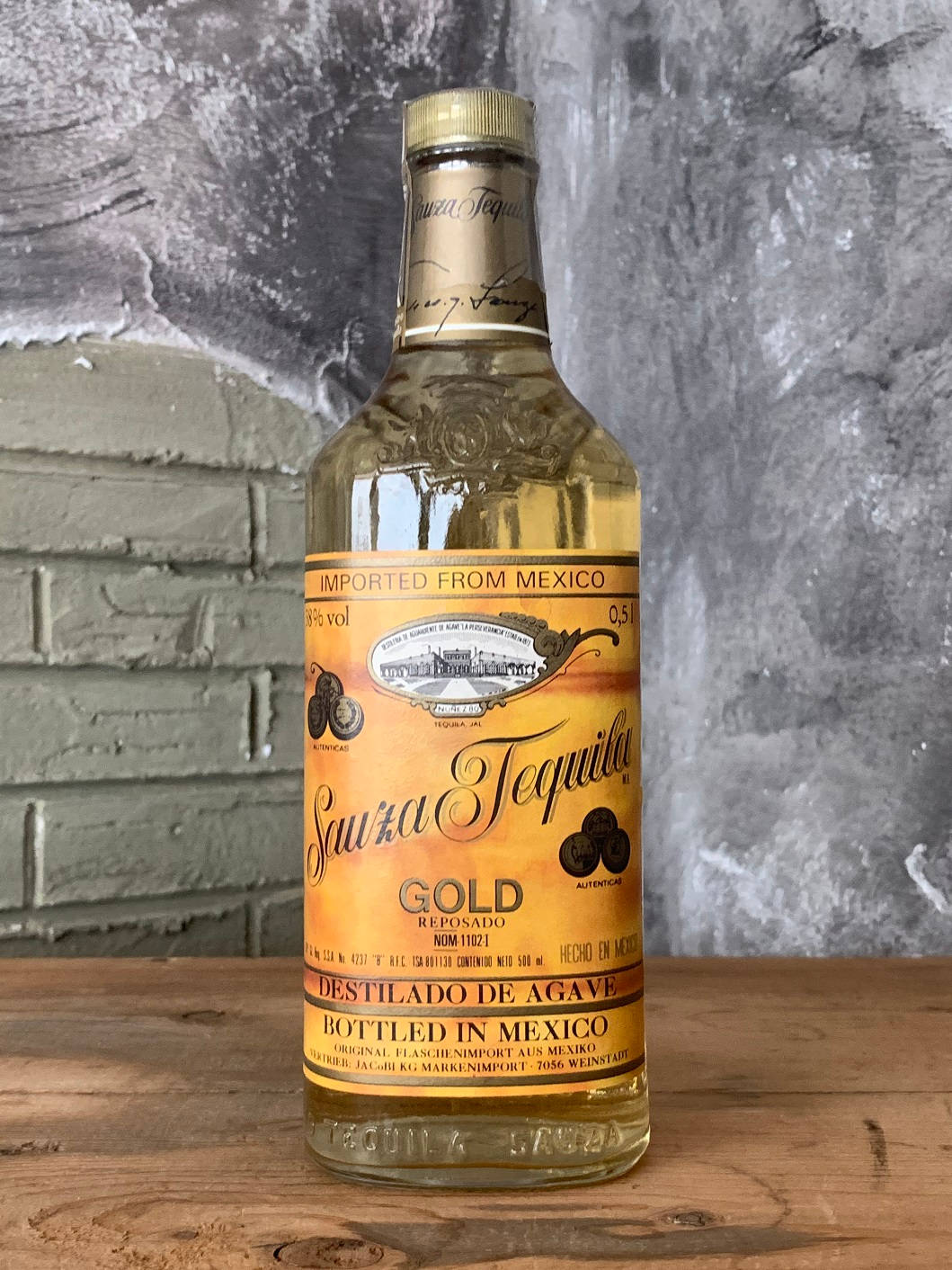 高清 桌布 壁紙 — 一瓶清澈的 Sauza Gold Tequila 放在質樸的木質表面上，背景是帶紋理的灰色磚牆。