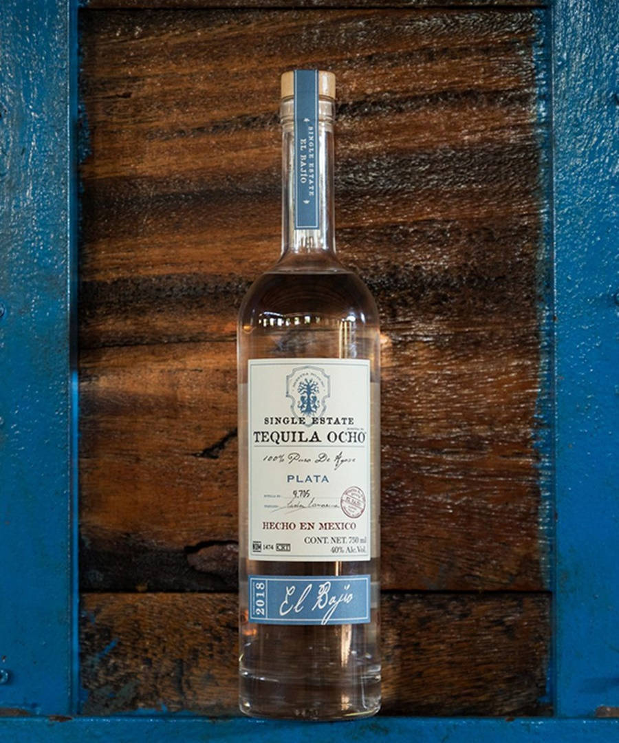 bottle 桌布 壁紙 — 一瓶清澈的 Single Estate Tequila Ocho Plata 置中於質樸的木質背景前，並以鮮豔的藍色油漆框起。