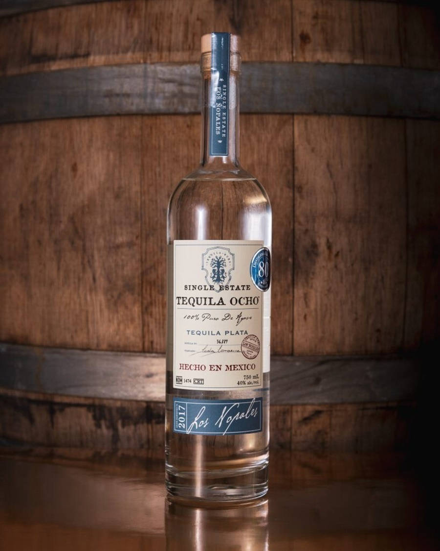bottle 桌布 壁紙 — 一瓶清澈的 Single Estate Tequila Ocho 酒瓶居中放在一個質樸的木桶前，帶有溫暖的環境燈光。