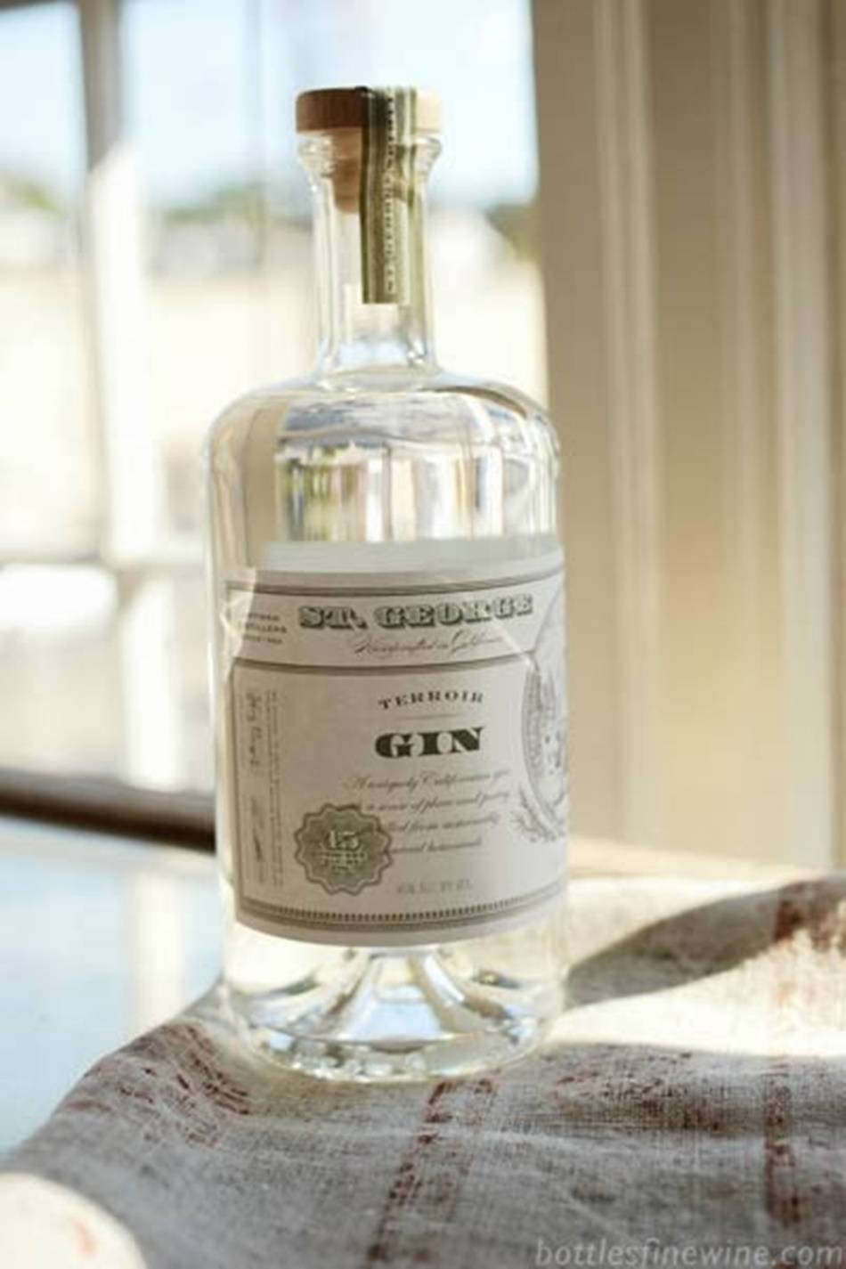 bottle 桌布 壁紙 — 一瓶清澈的 St. George Terroir Gin 放在有圖案的布上，靠近陽光照射的窗戶，光線柔和自然。
