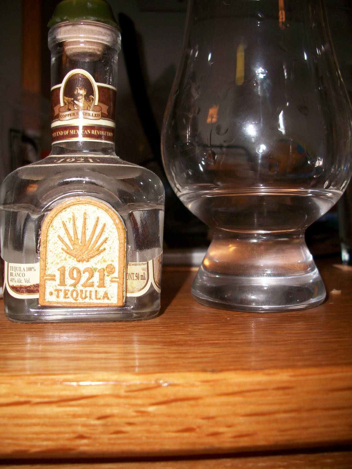 drinks 桌布 壁紙 — 一瓶清澈的1921 Tequila Blanco放在溫暖的木質表面上，旁邊有一個空玻璃杯。