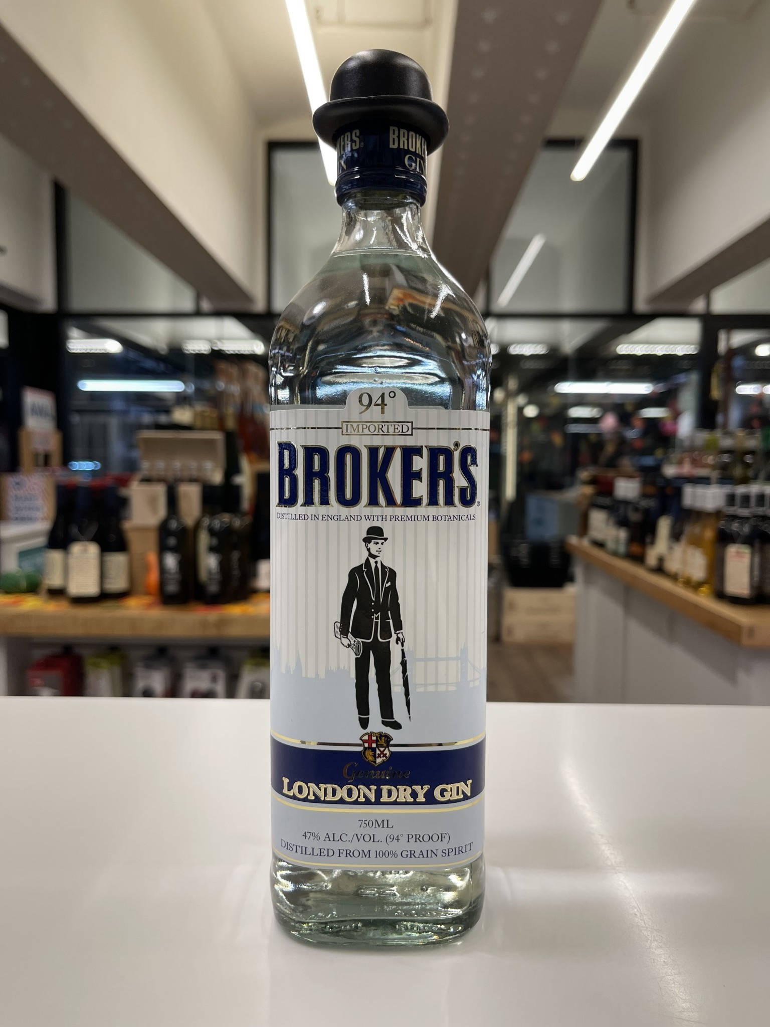 bottle 桌布 壁紙 — 一瓶清澈的Broker's London Dry Gin酒瓶放在白色檯面上，背景是光線充足的酒舖，貨架上擺滿了酒瓶。