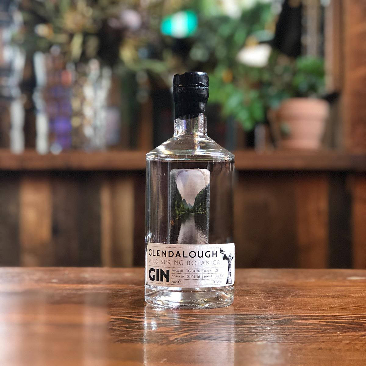 drinks 桌布 壁紙 — 一瓶清澈的Glendalough Wild Spring Botanical Gin放在拋光木吧檯上，背景是模糊的綠植和架子。