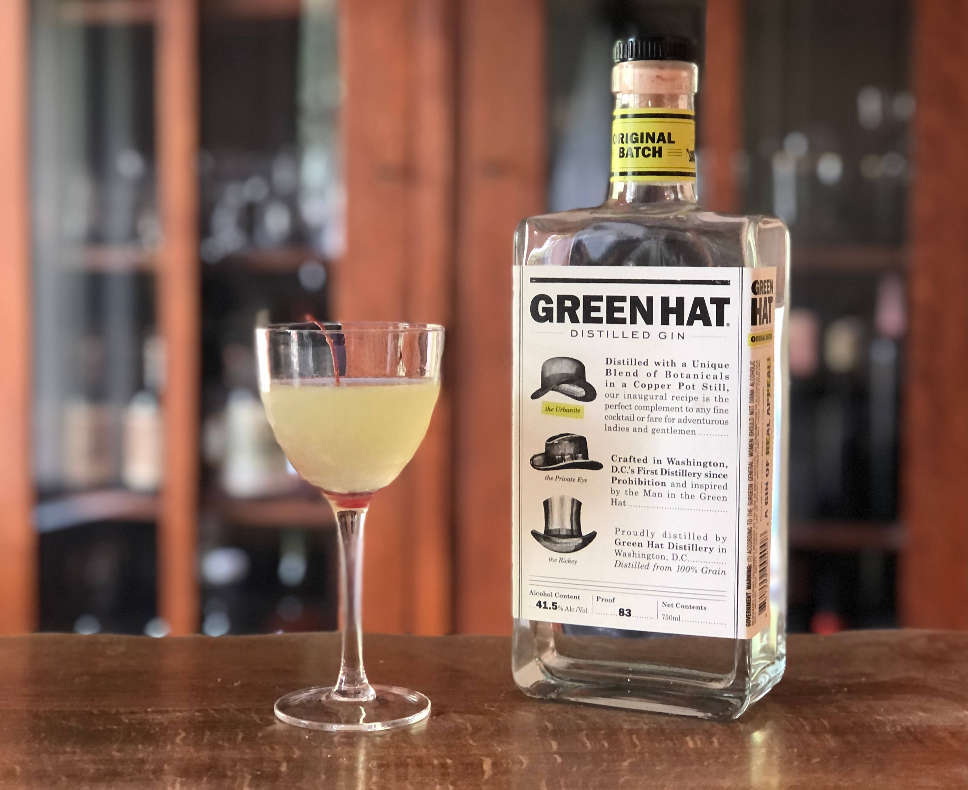 alcohol 桌布 壁紙 — 一瓶清澈的Green Hat蒸餾琴酒放在木製吧檯上，旁邊是一杯淡黃色雞尾酒，盛在庫佩杯中，背景有模糊的層架。
