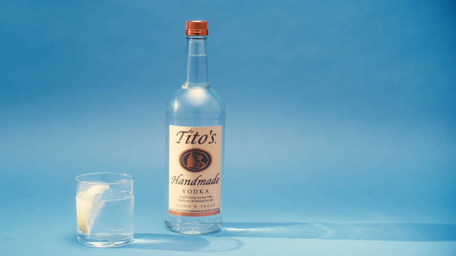Lemon 桌布 壁紙 — 一瓶清澈的Tito's Handmade Vodka放在一個裝有冰塊和檸檬角的玻璃杯旁邊，背景為純藍色。
