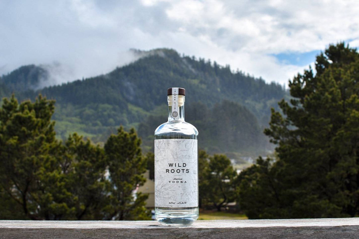 bottle 桌布 壁紙 — 一瓶清澈的Wild Roots伏特加放在木欄杆上，背景是霧氣瀰漫的綠色山脈和松樹。