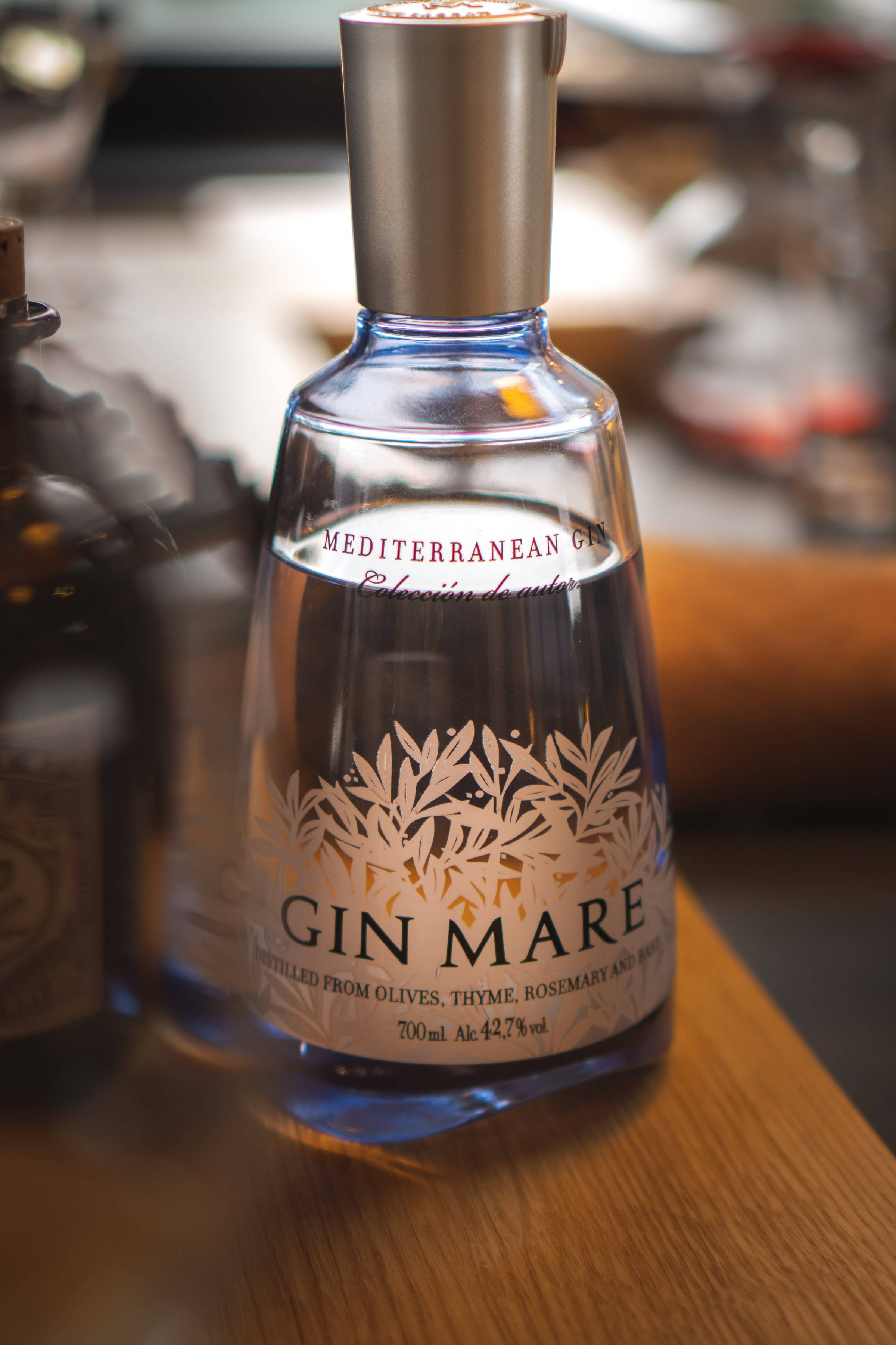 drinks 桌布 壁紙 — 一瓶清澈的玻璃瓶 Gin Mare 地中海琴酒放置在木質表面上，帶有柔和溫暖的燈光和模糊的背景元素。