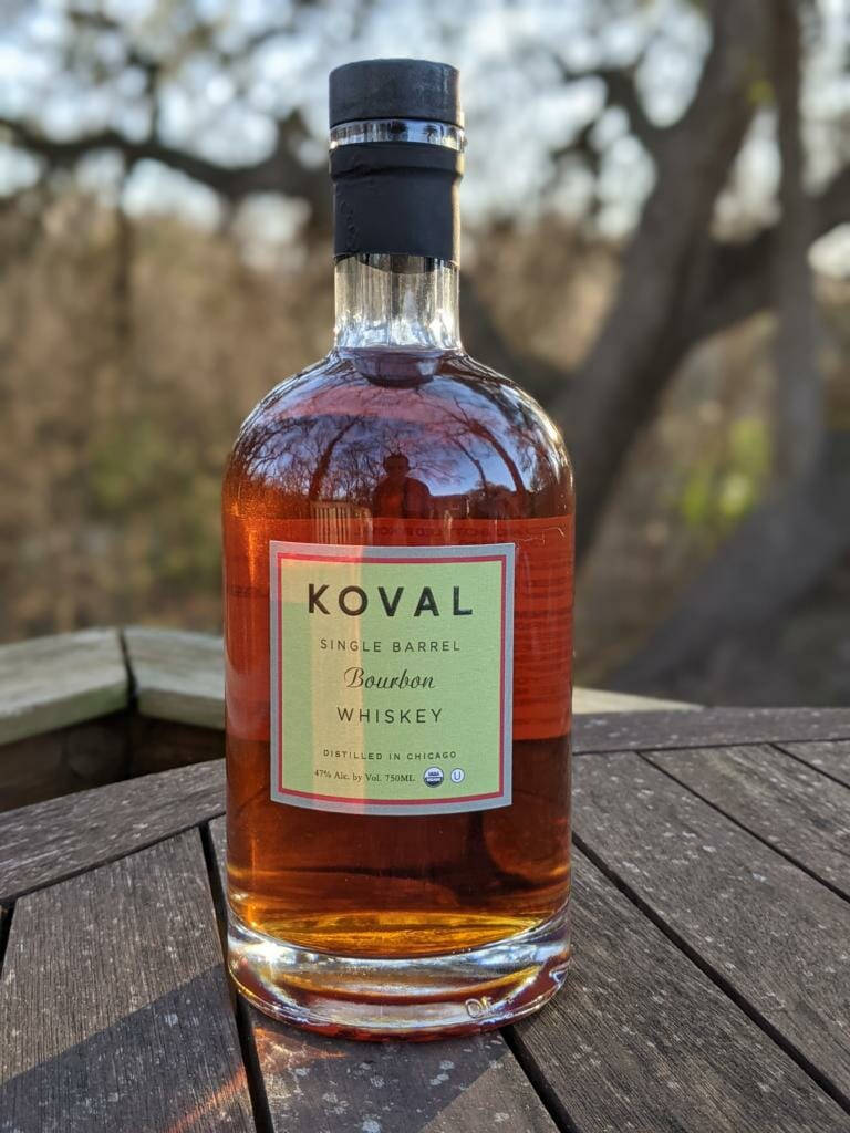 alcohol 桌布 壁紙 — 一瓶清澈的玻璃瓶裝Koval單桶波本威士忌放在戶外一張風化的木桌上，背景是模糊的樹木。