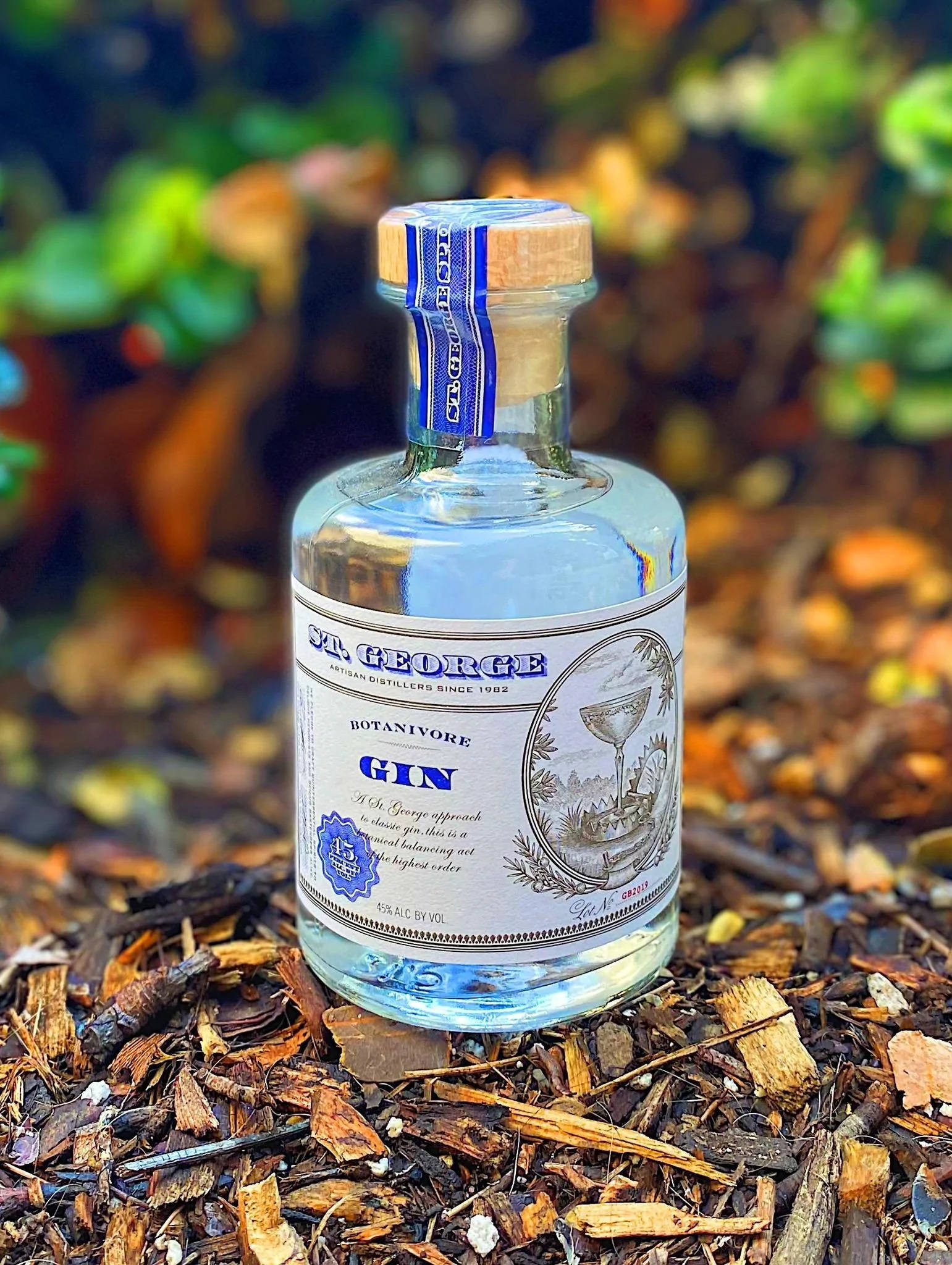 bottle 桌布 壁紙 — 一瓶清澈的玻璃瓶裝聖喬治植物學家琴酒放在木屑覆蓋物上，背景是模糊的綠色樹葉。