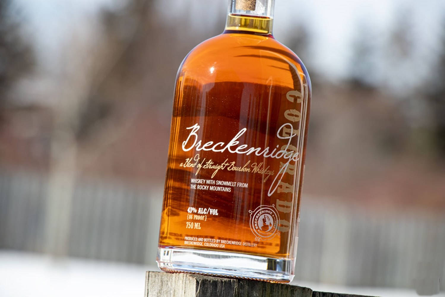 Drink 桌布 壁紙 — 一瓶清澈的琥珀色 Breckenridge 波本威士忌放在木柱上，背景是白雪皚皚的戶外景色。