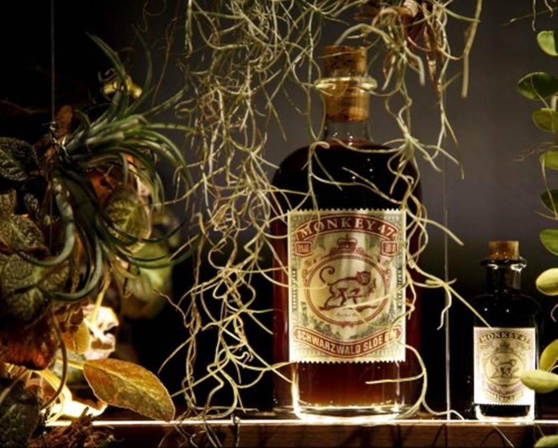 cocktail 桌布 壁紙 — 一瓶溫暖照亮的Monkey 47 Schwarzwald Sloe Gin優雅地放在木架上，周圍環繞著茂密的綠色植物和精緻的空氣植物。