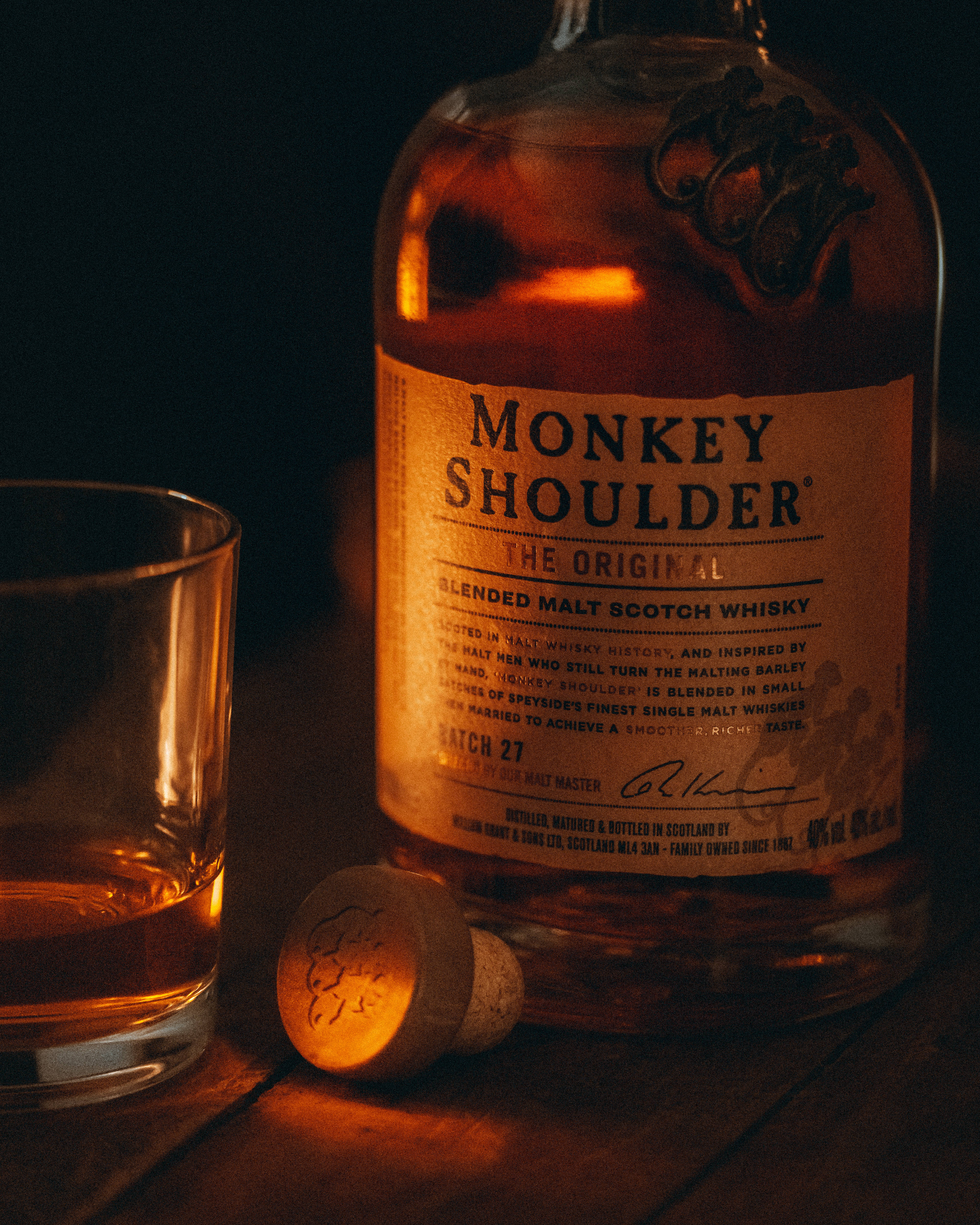 alcohol 桌布 壁紙 — 一瓶溫暖燈光照亮的Monkey Shoulder威士忌，旁邊放著一個玻璃杯和軟木塞，在昏暗的環境光線下置於木桌上。